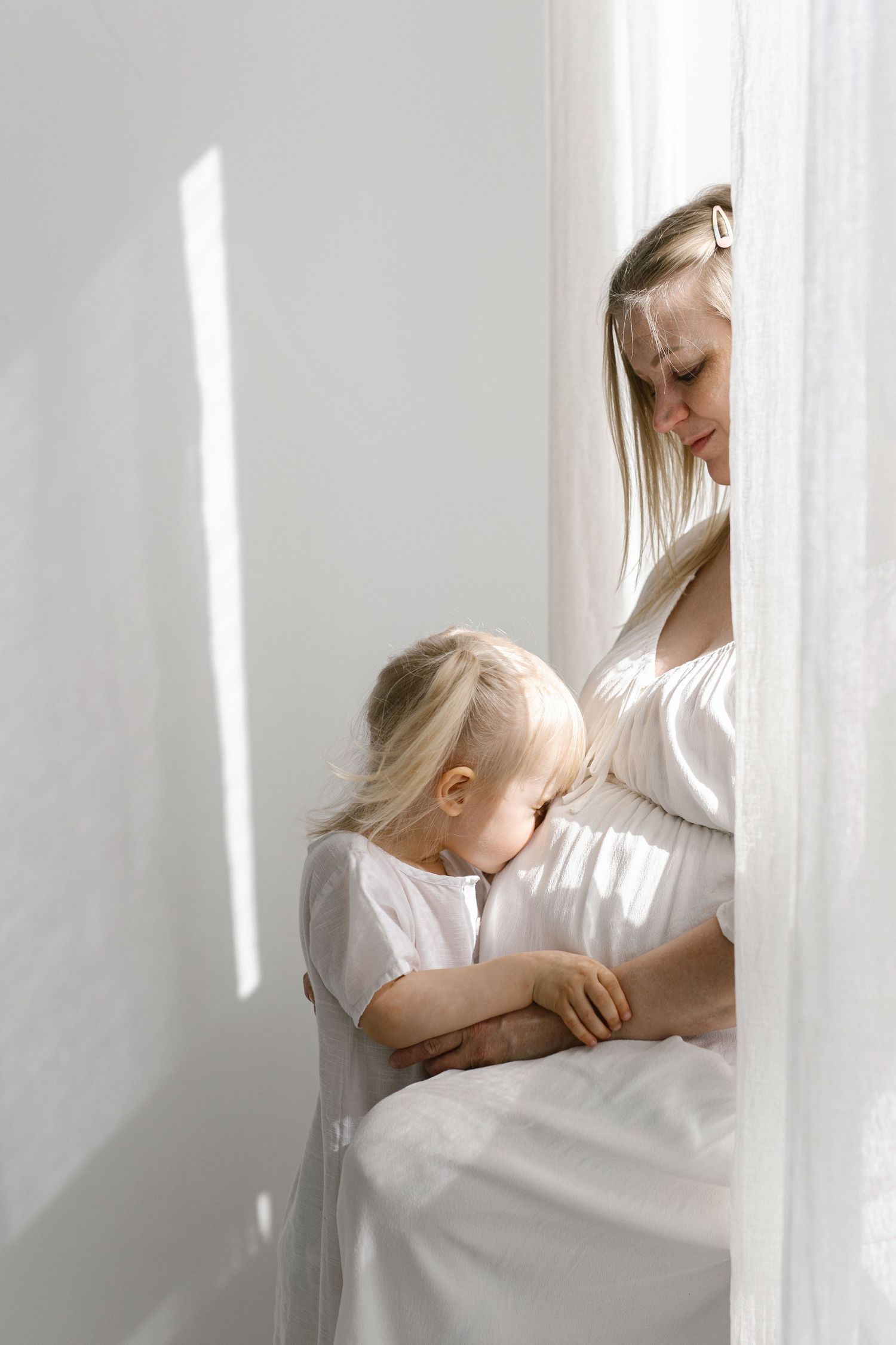 Pregnant woman in white dress tenderly bonding with her young blonde toddler by a sunlit window.