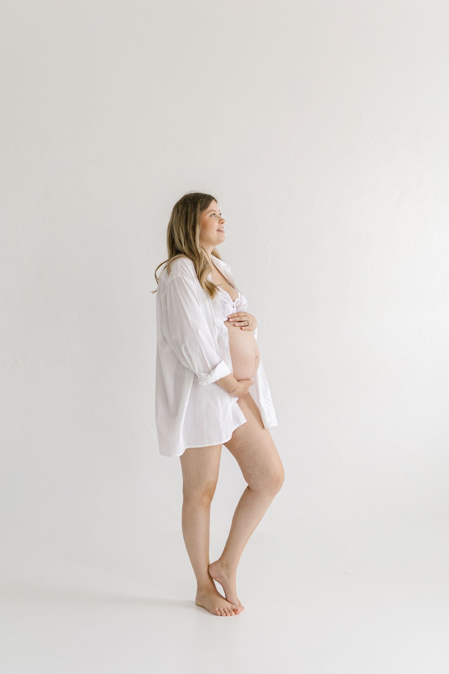 Pregnant woman in white shirt and nude bra cradling baby bump, standing barefoot against white background.