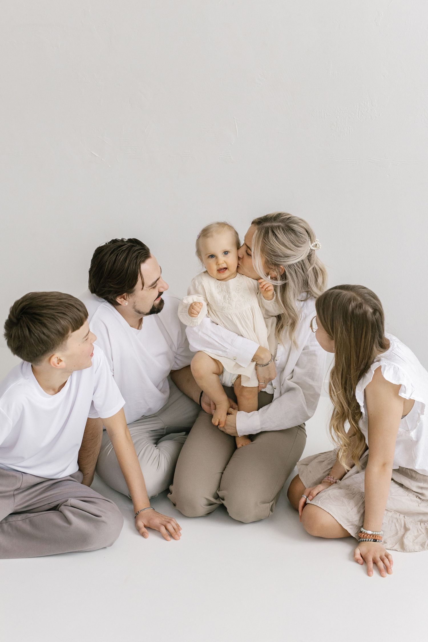 Family of five sitting together on white floor, parents kissing baby while two older children look on lovingly.