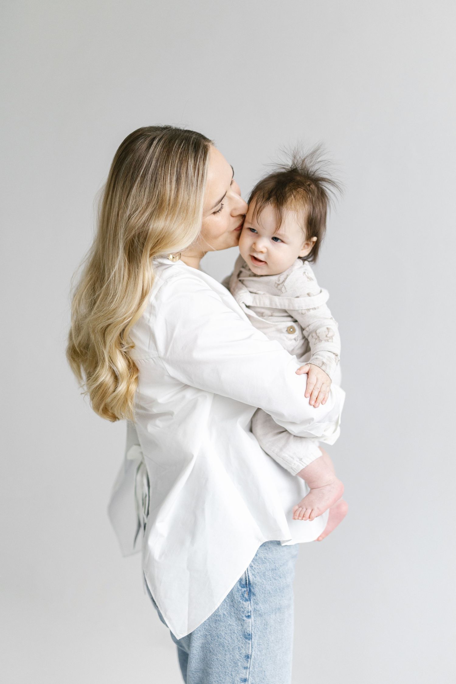 A blonde mother in a white shirt and jeans kisses her smiling baby girl in a light gray studio setting.