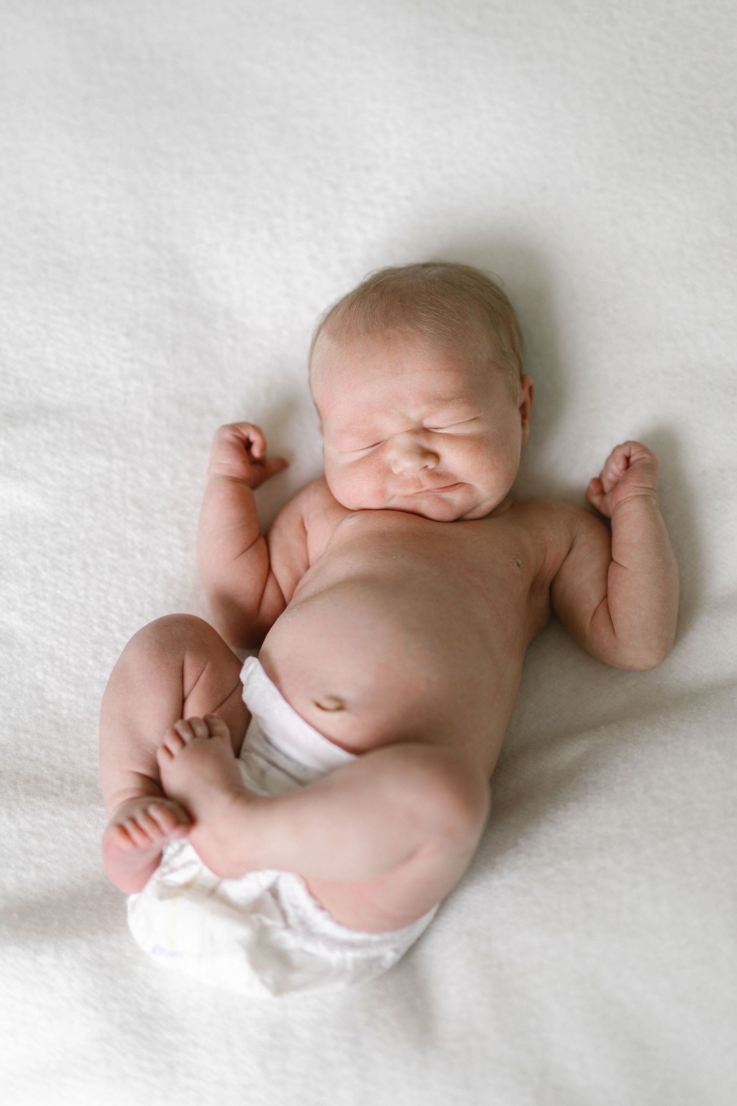 Newborn baby in white diaper lying on back on white blanket, scrunching face with arms raised.