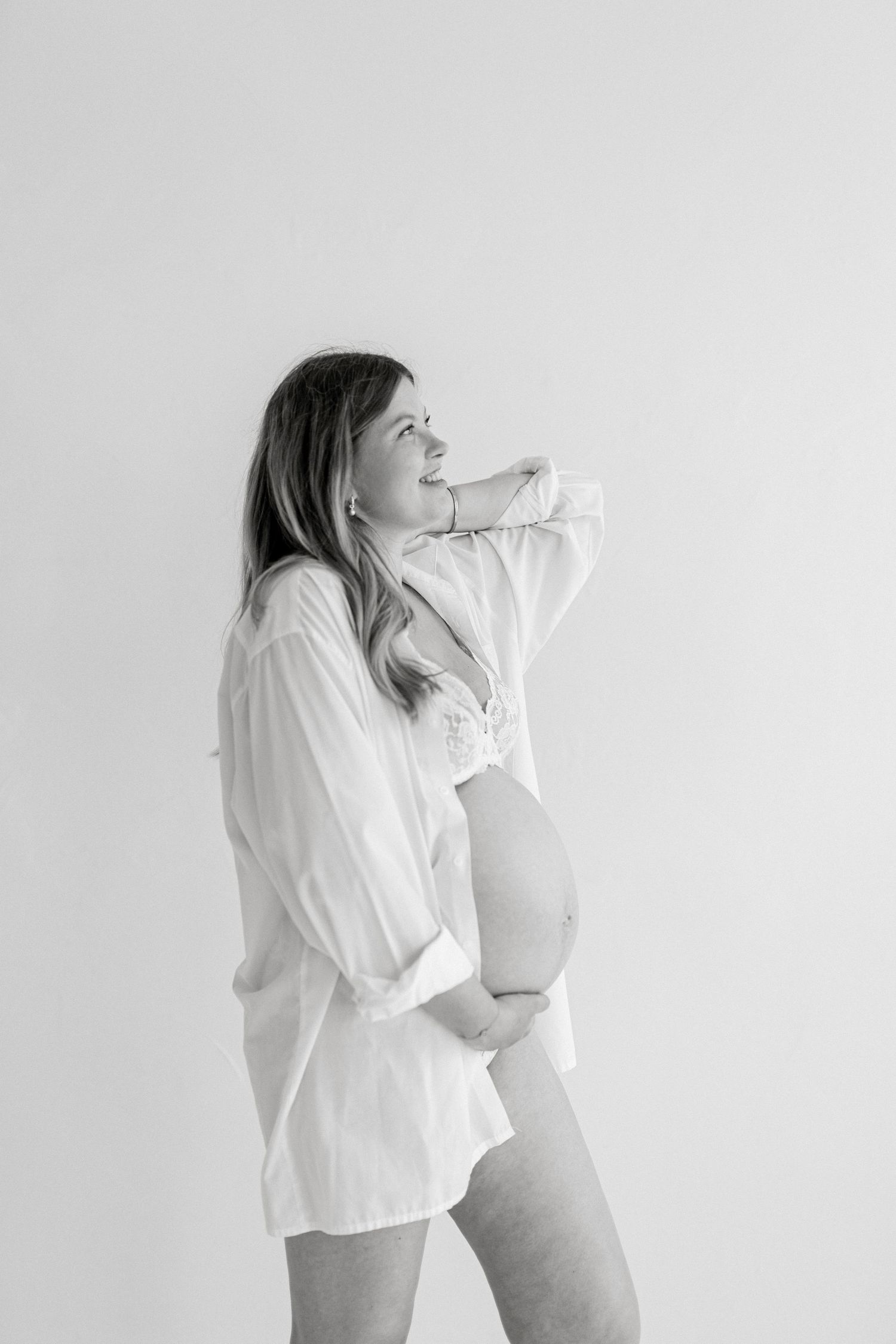 Black and white maternity photo of a woman in an open white shirt cradling her baby bump while looking up.