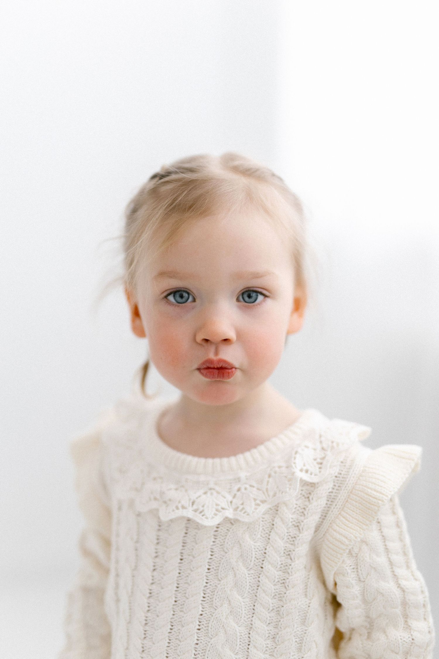 A young toddler girl with blue eyes wearing a white cable-knit sweater poses for a portrait photo.