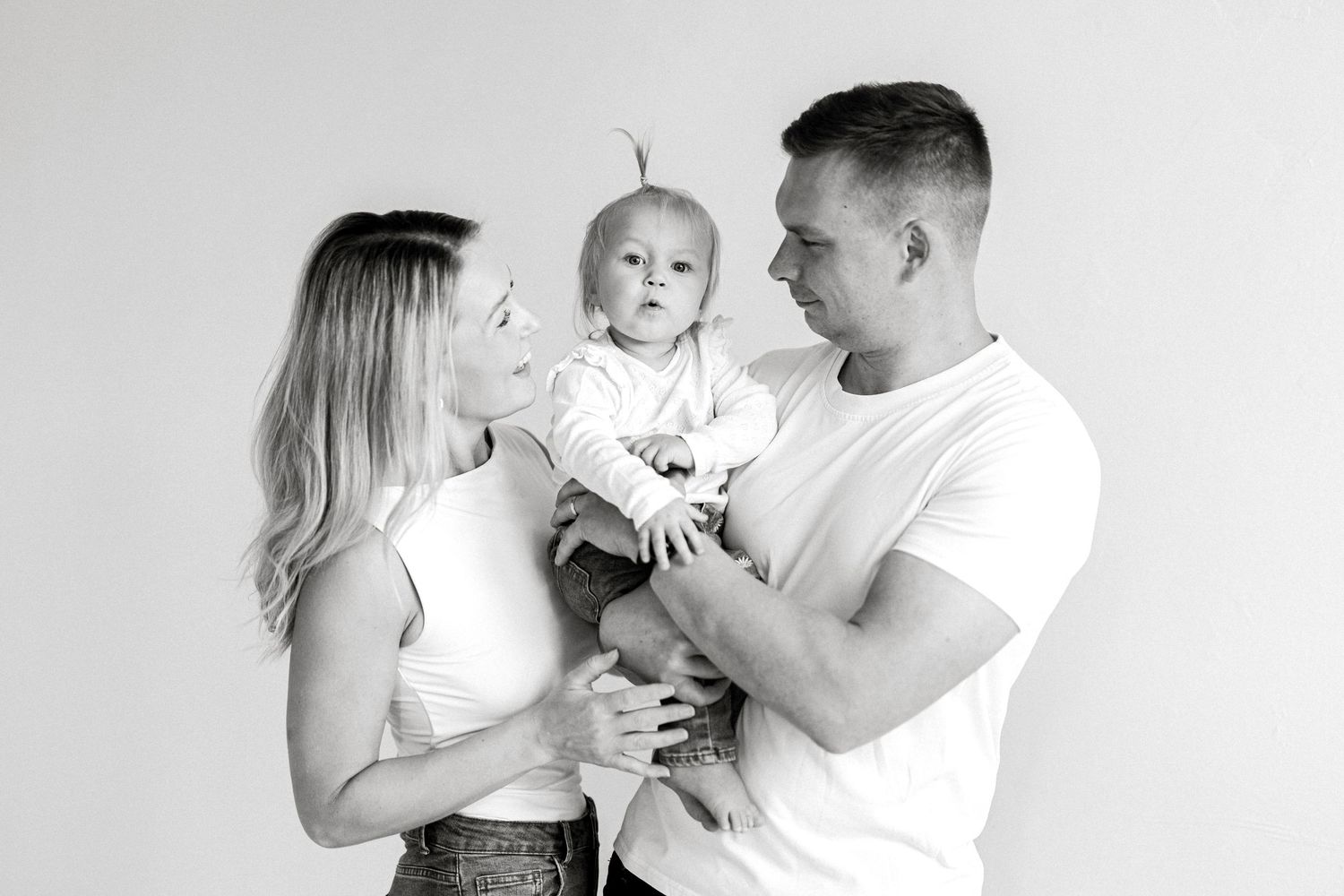 Black and white family portrait of parents smiling at their baby in a bright studio setting.
