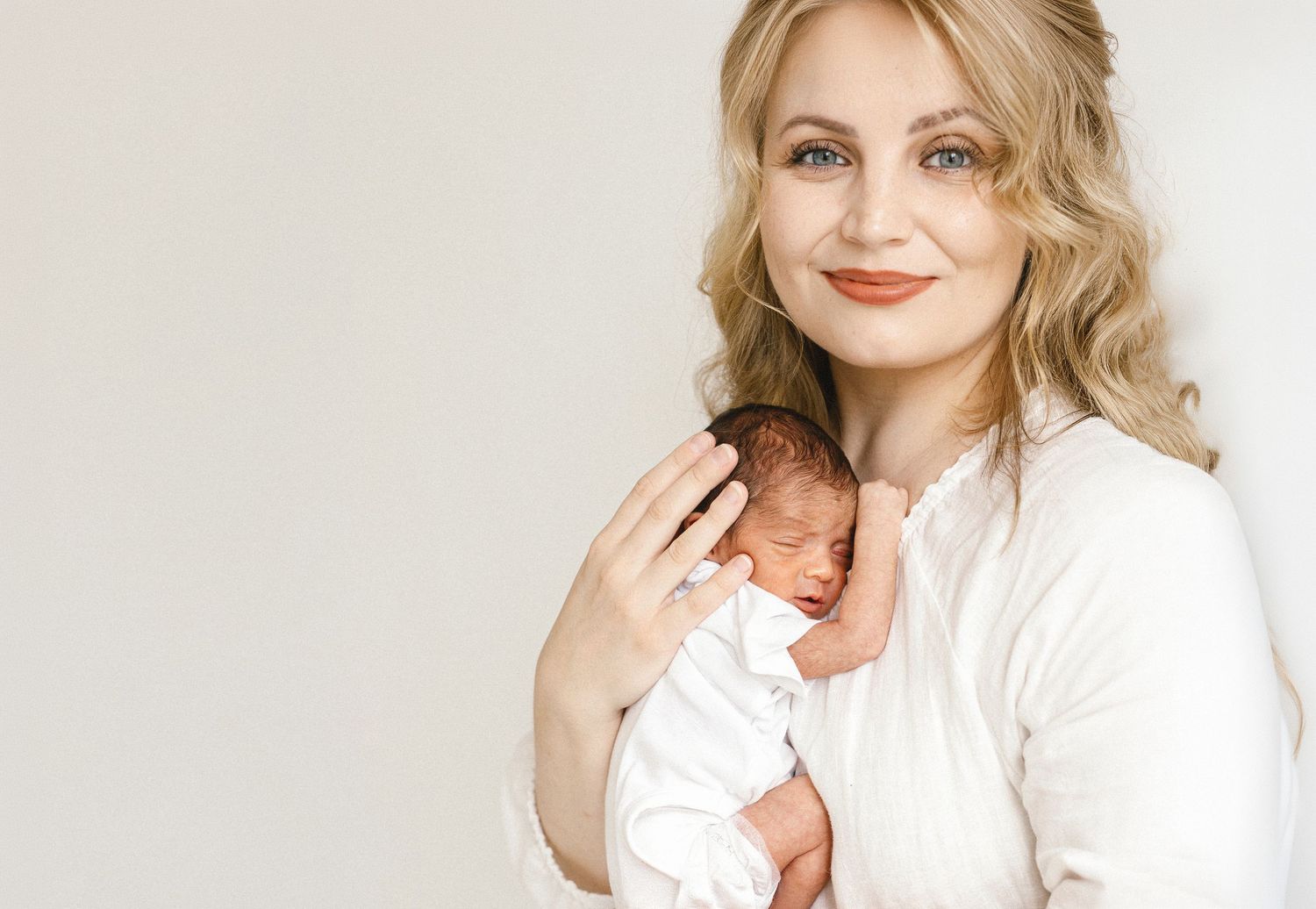 Smiling blonde woman in white blouse gently holding a sleeping newborn baby against a light background.