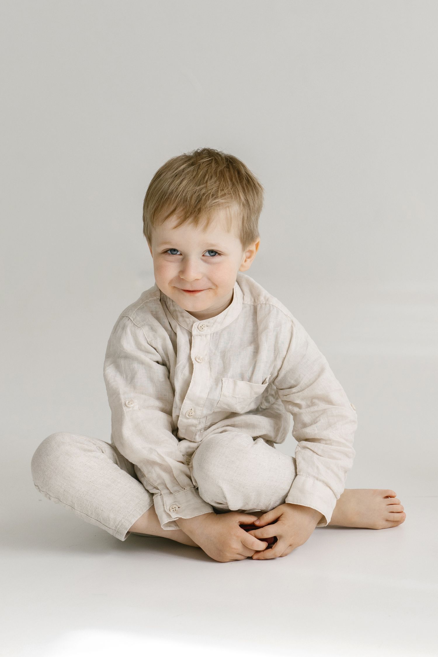 Young child in linen outfit sitting cross-legged on white background with gentle smile.