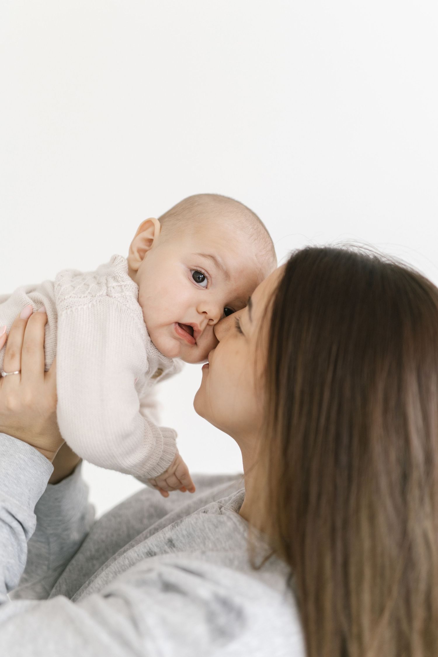 A loving mother gently kisses her adorable baby girl dressed in pink, against a white background.