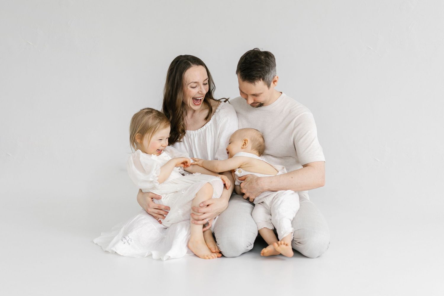 A happy family of four in white outfits sits together on a white background, parents lovingly holding their newborn and toddler.