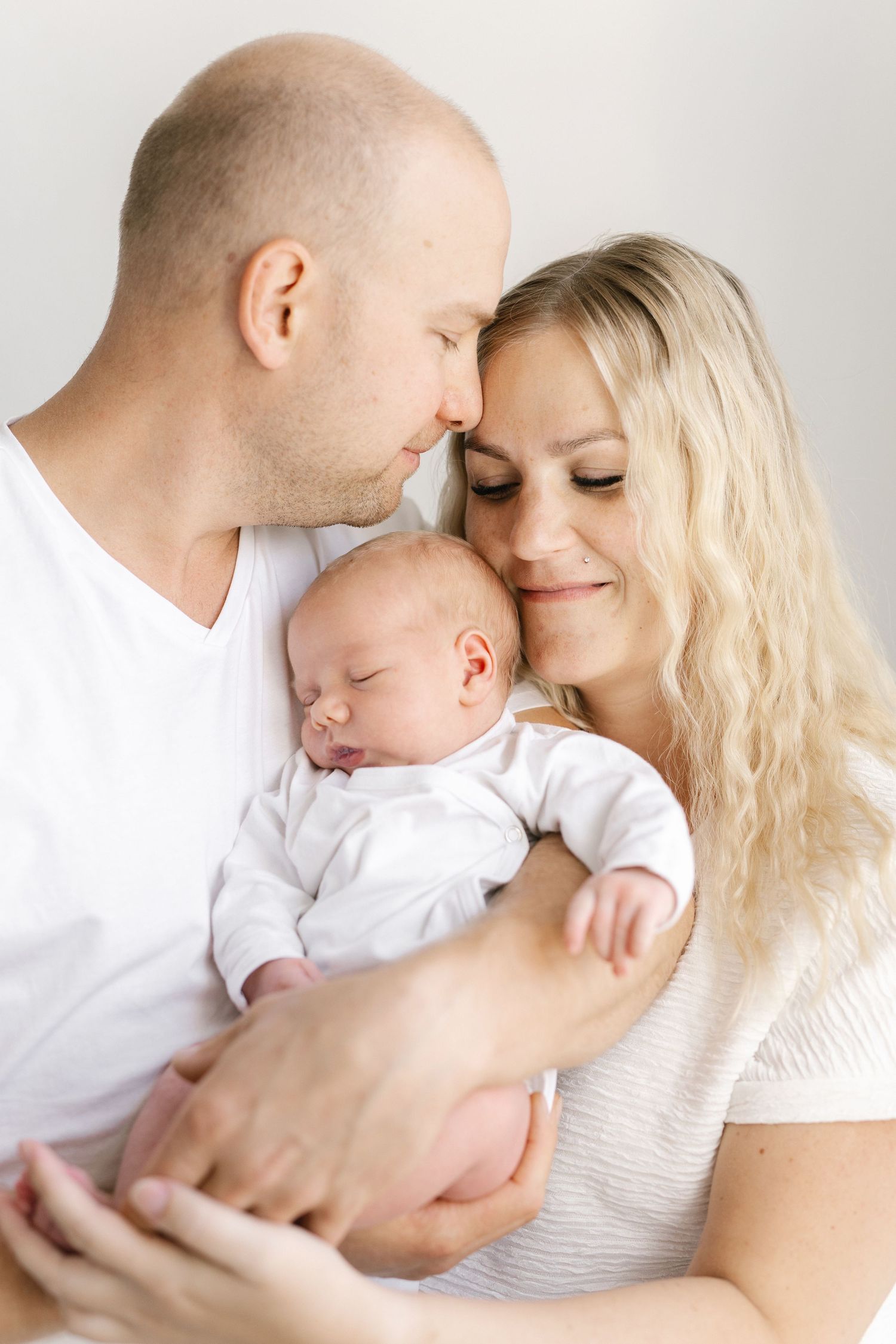 A loving couple in white clothing tenderly holds their sleeping newborn baby, touching foreheads in an intimate family portrait.