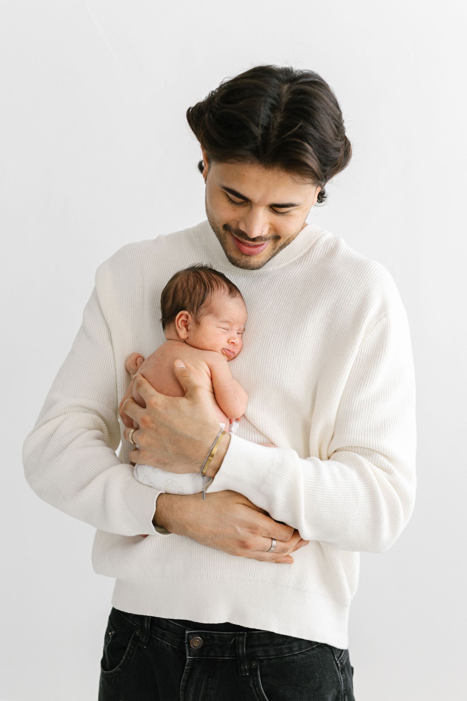 Smiling young father in white sweater gently cradling sleeping newborn baby against light background.