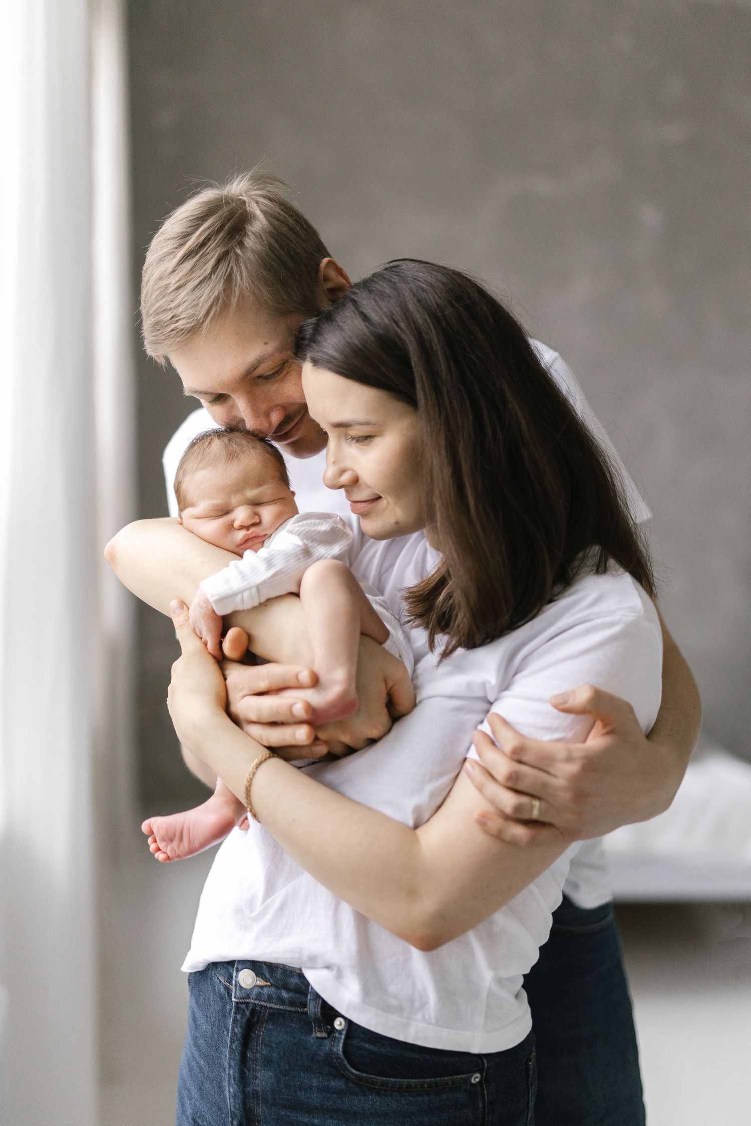 Two parents in white shirts tenderly hold their sleeping newborn baby together in a bright modern room.