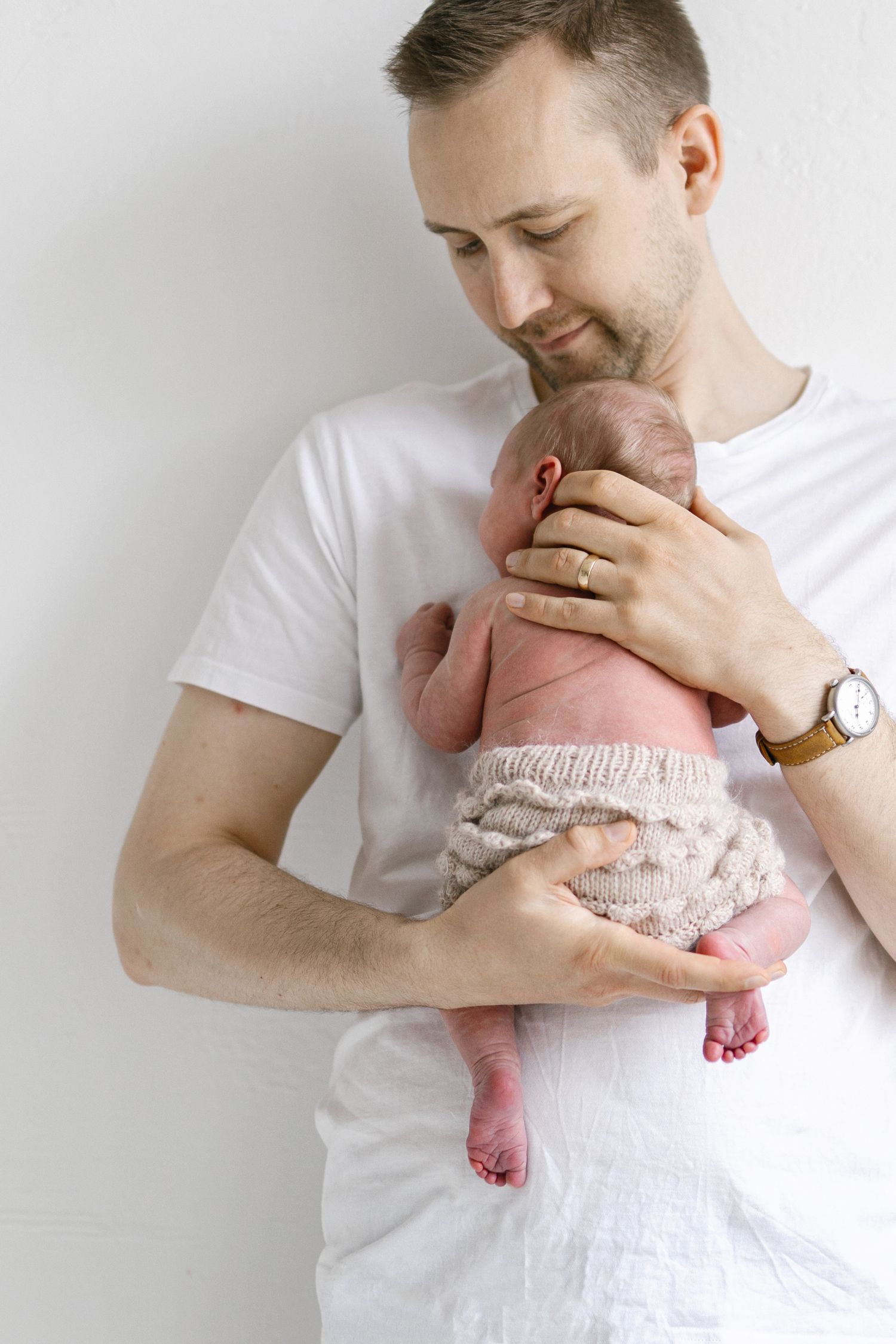 A father in a white t-shirt tenderly holds his newborn baby dressed in a knit wrap against a white background.