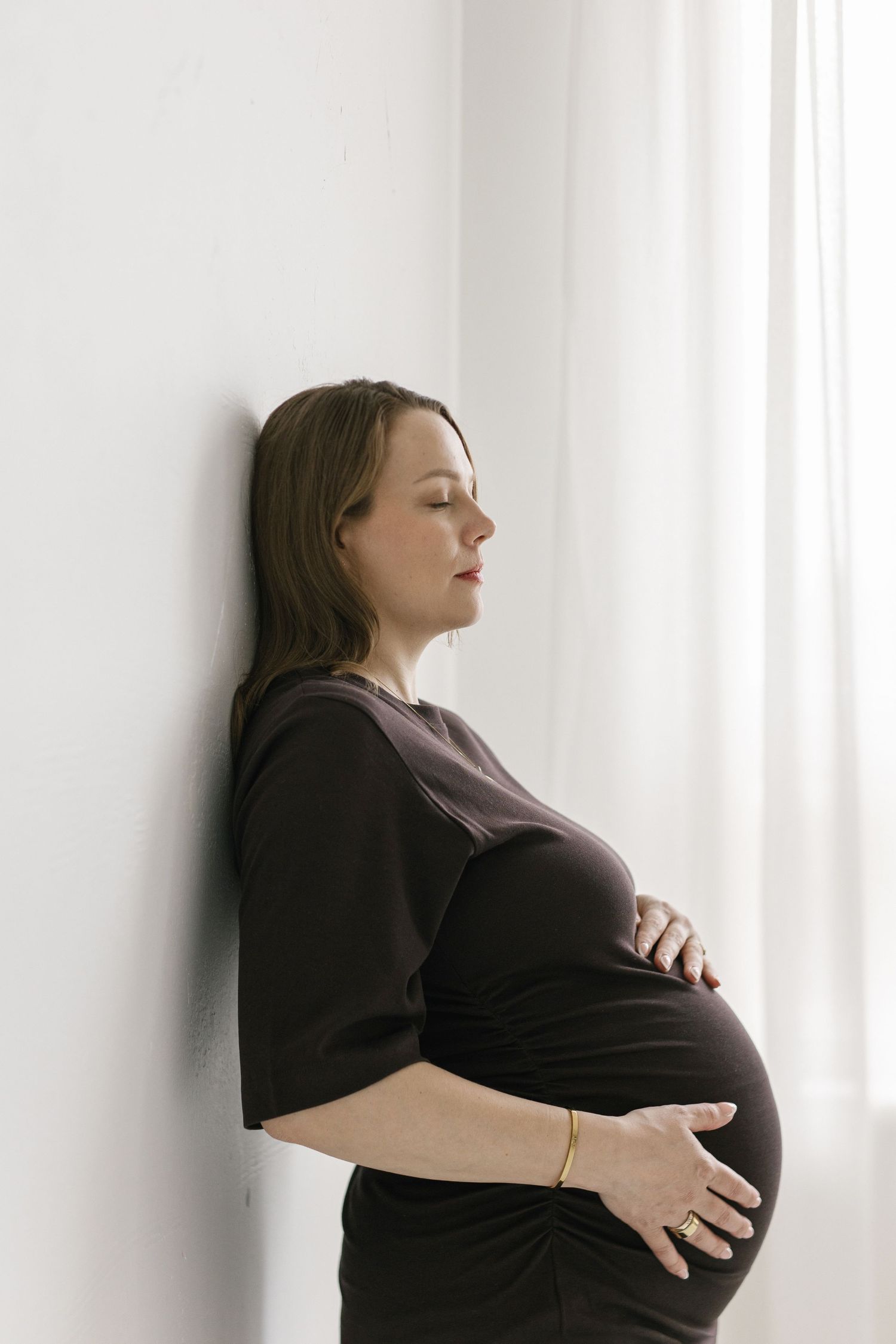 Pregnant woman in black dress leaning against white wall with eyes closed, cradling her baby bump.