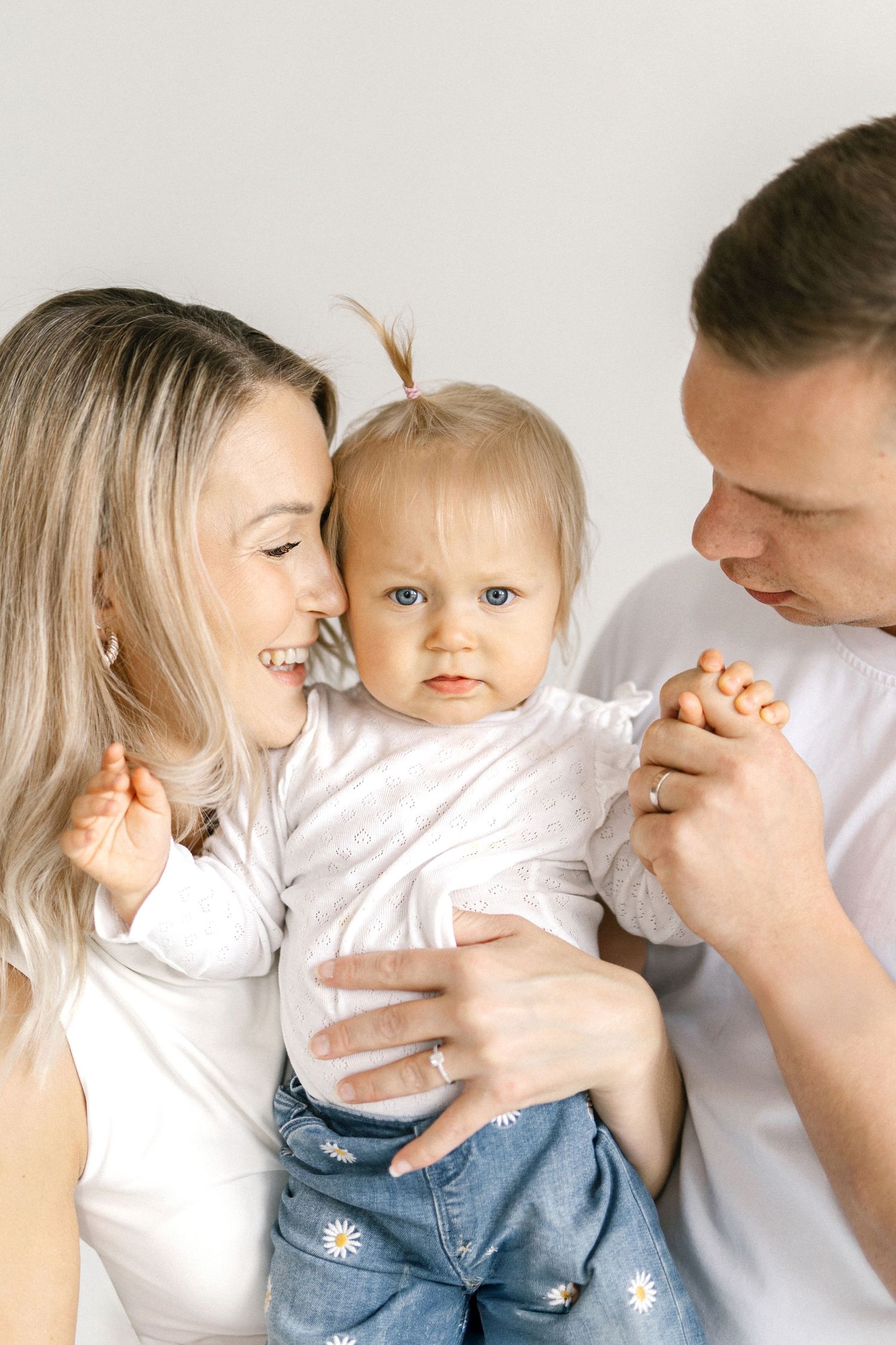 Happy young family with blonde mom and dad in white shirts holding their baby girl with blue eyes and a tiny ponytail.