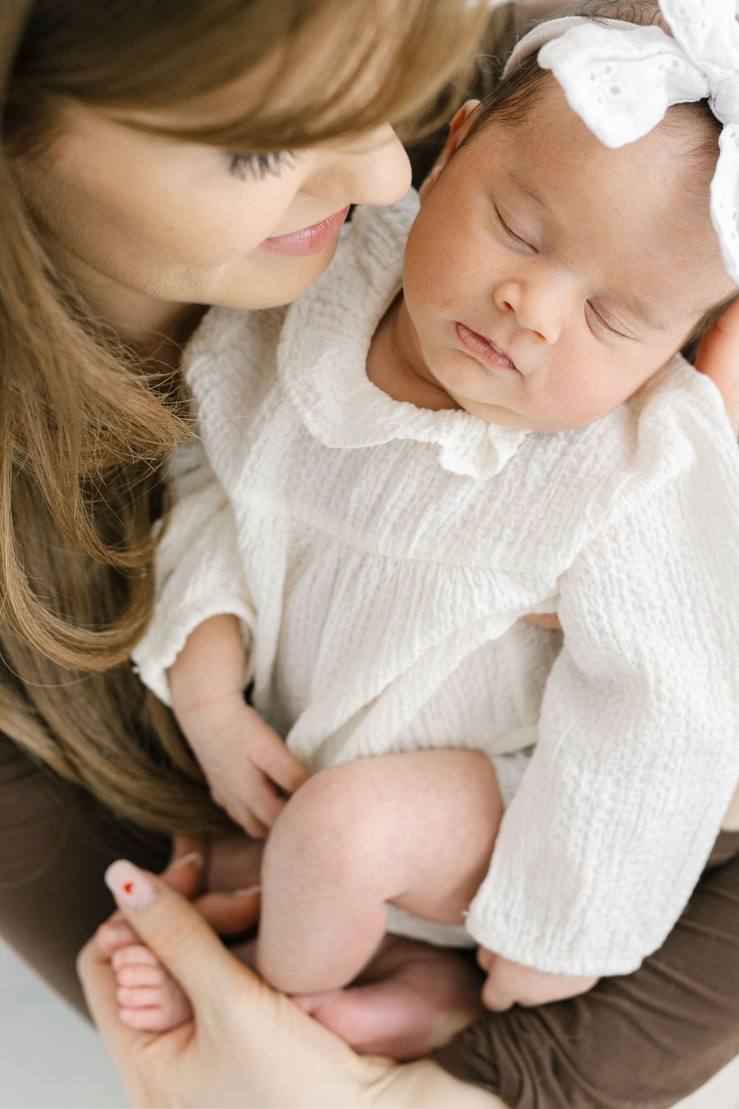 A mother tenderly holds her sleeping newborn baby girl dressed in white with a bow headband.