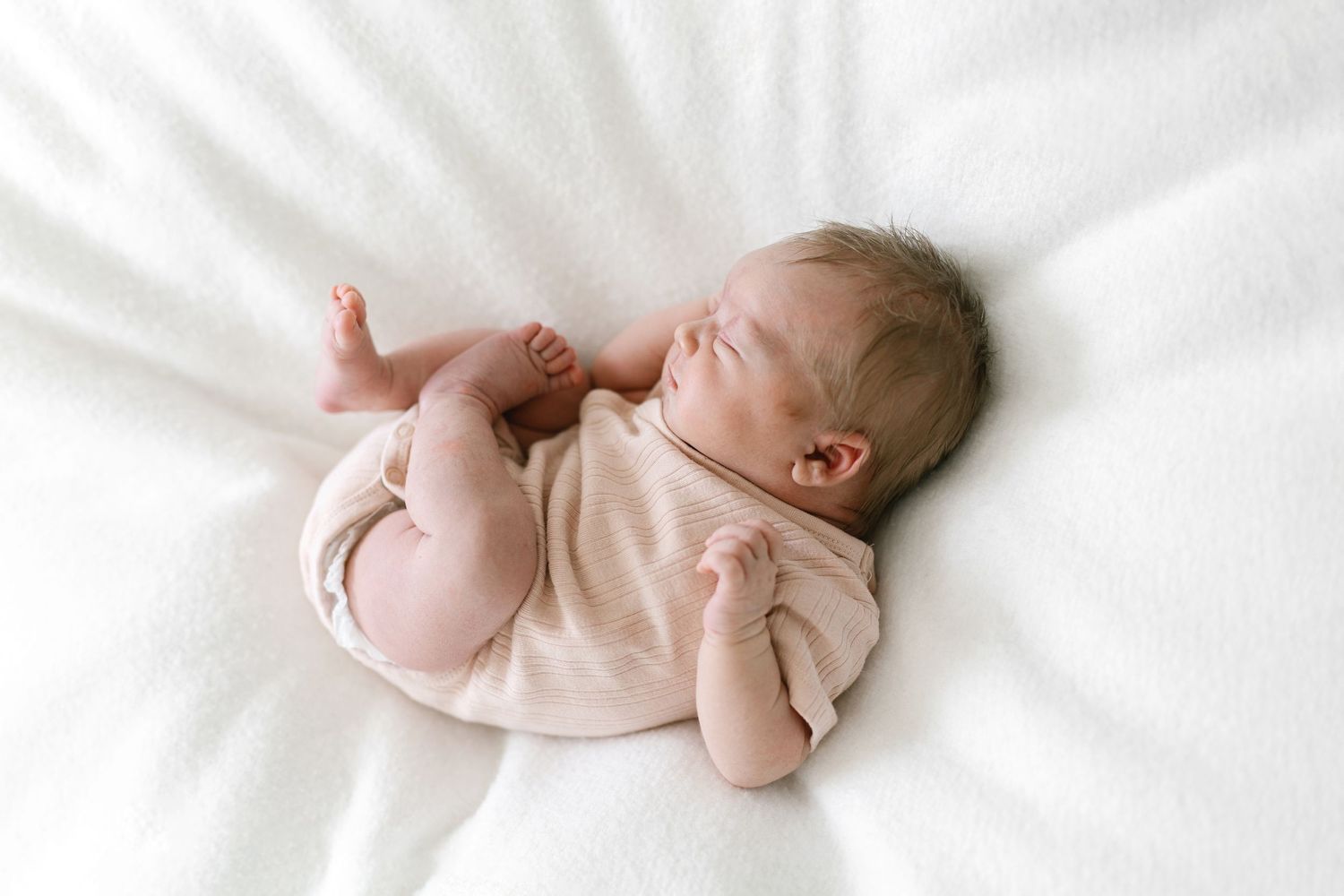 Newborn baby sleeping peacefully on white fluffy blanket wearing a soft beige onesie.
