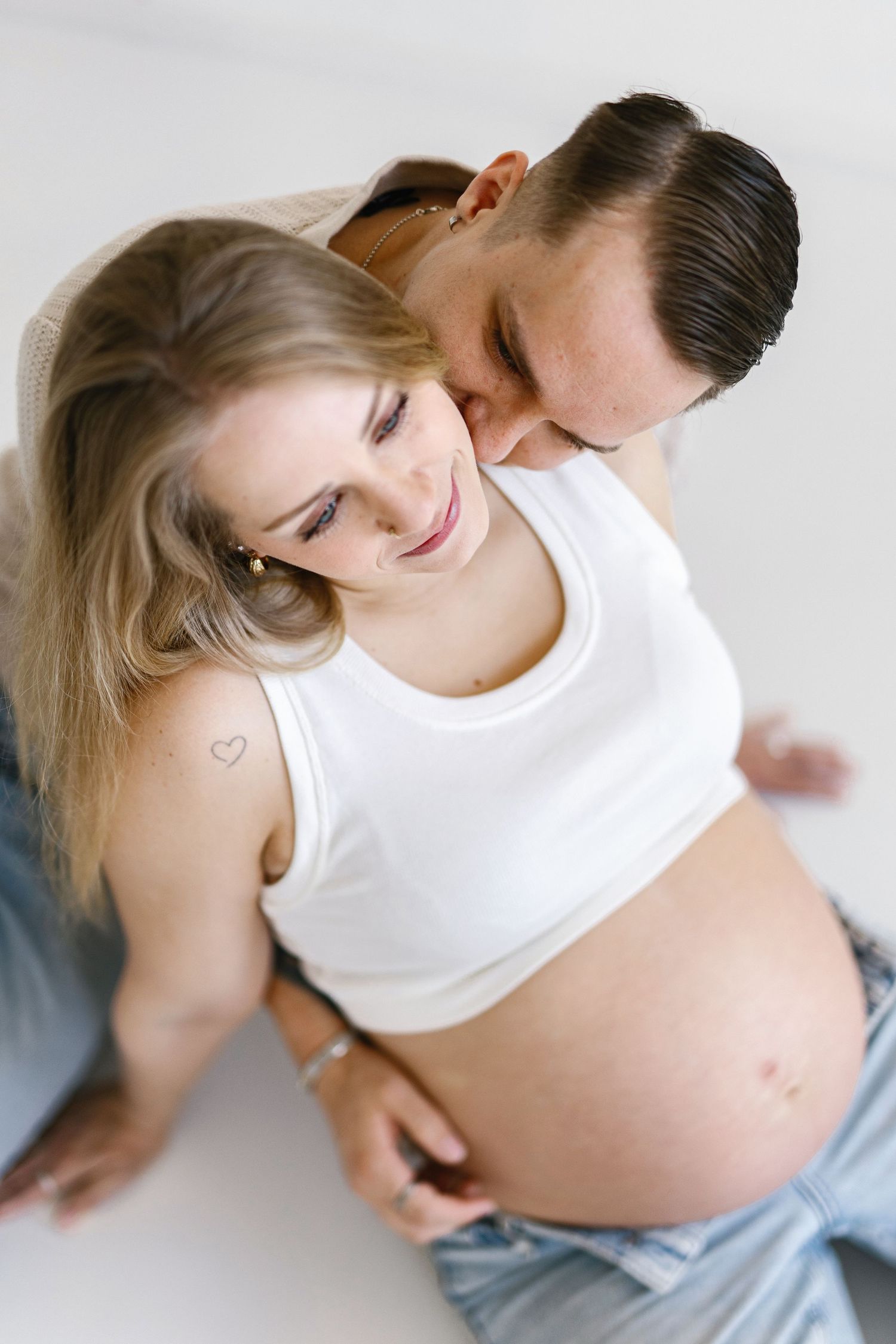Pregnant woman in white crop top smiles as partner kisses her head while tenderly holding her baby bump.