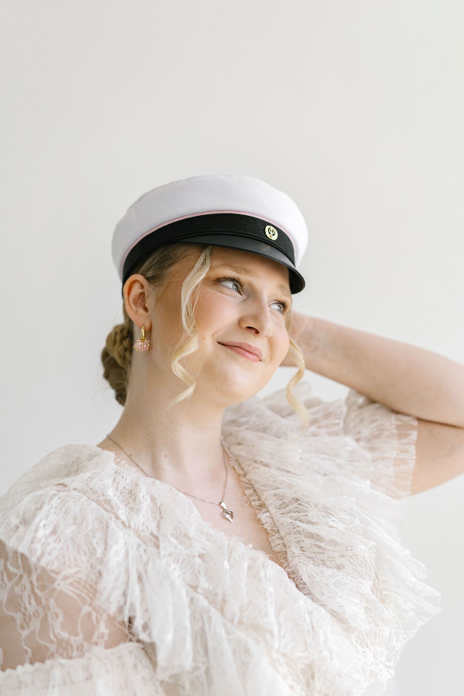 Blonde woman in white lace dress and sailor cap looking up, holding hat with hand against light background.