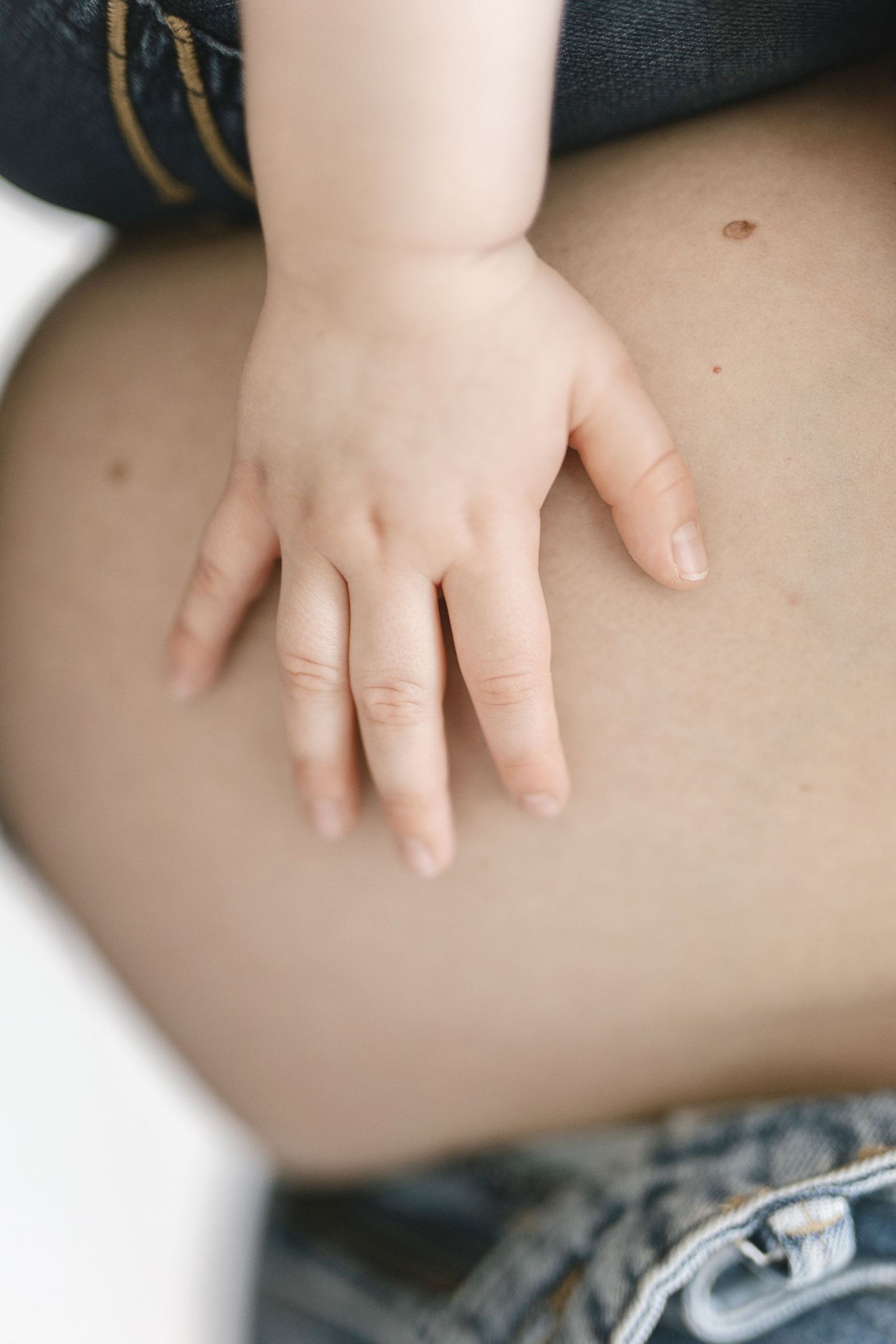 A small baby hand resting gently on a mother's bare pregnant belly above denim jeans.