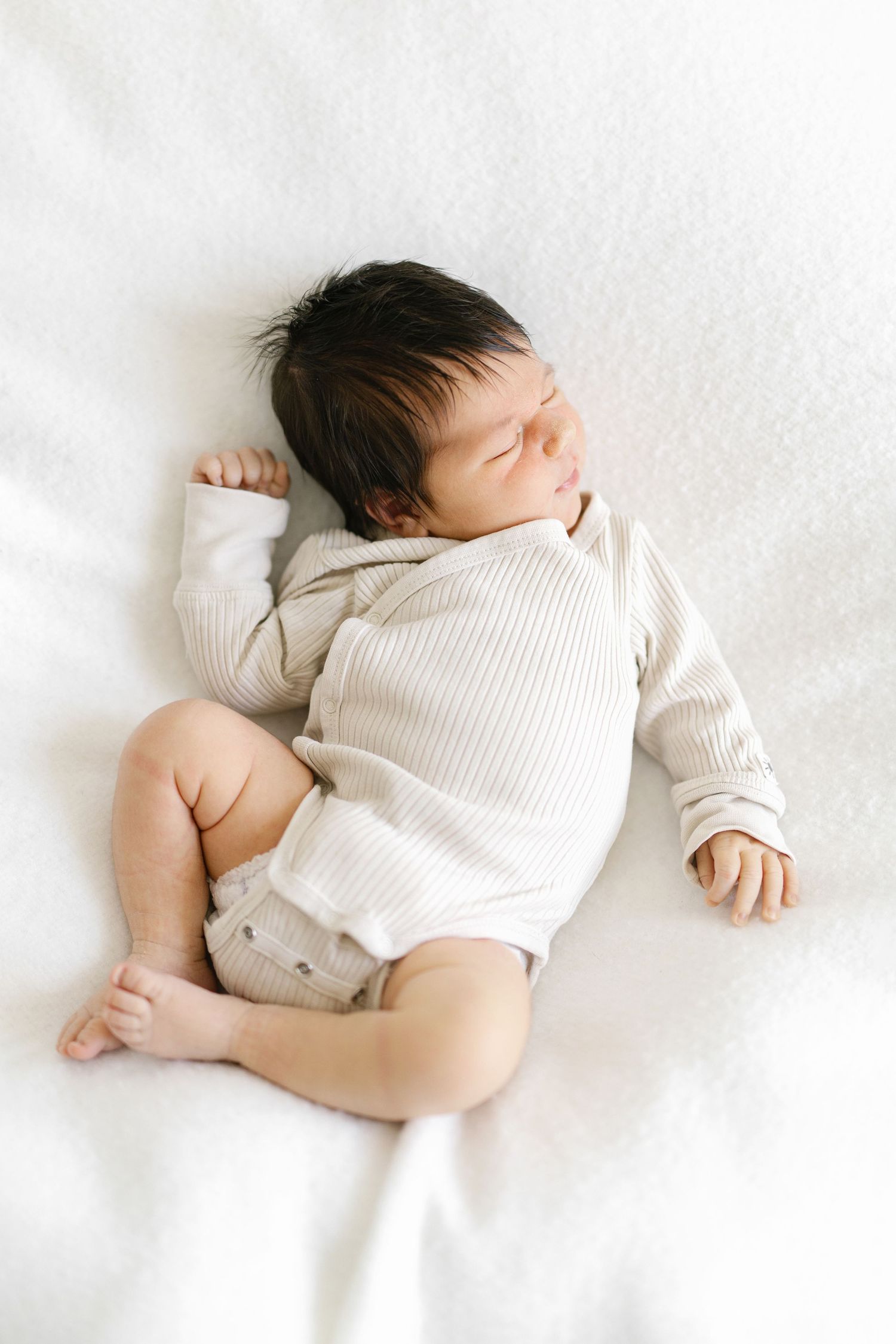 A peaceful newborn baby sleeping on a white blanket wearing a ribbed beige onesie.