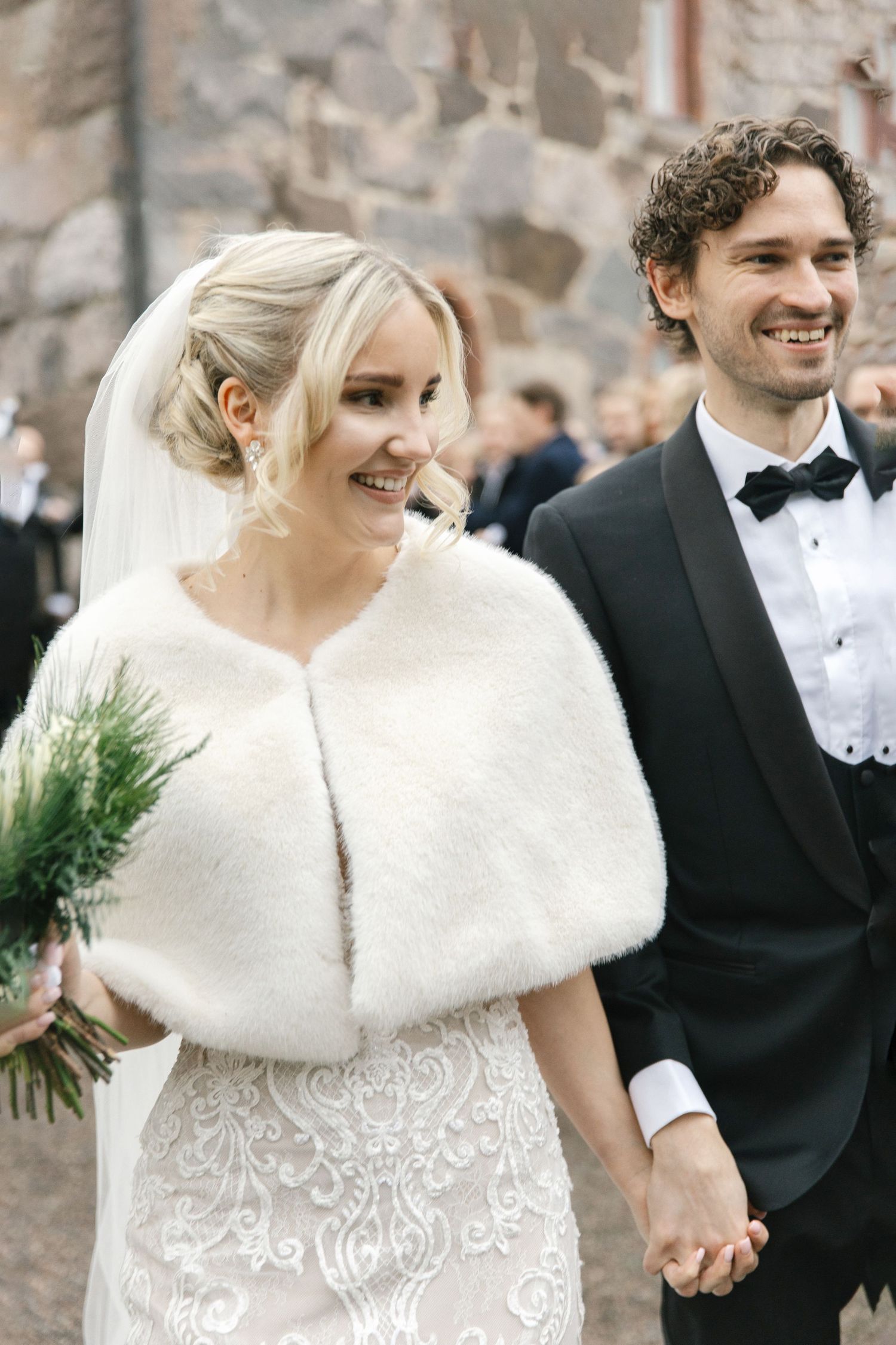 Winter wedding couple in formal attire with bride wearing white fur wrap and lace dress against stone wall.