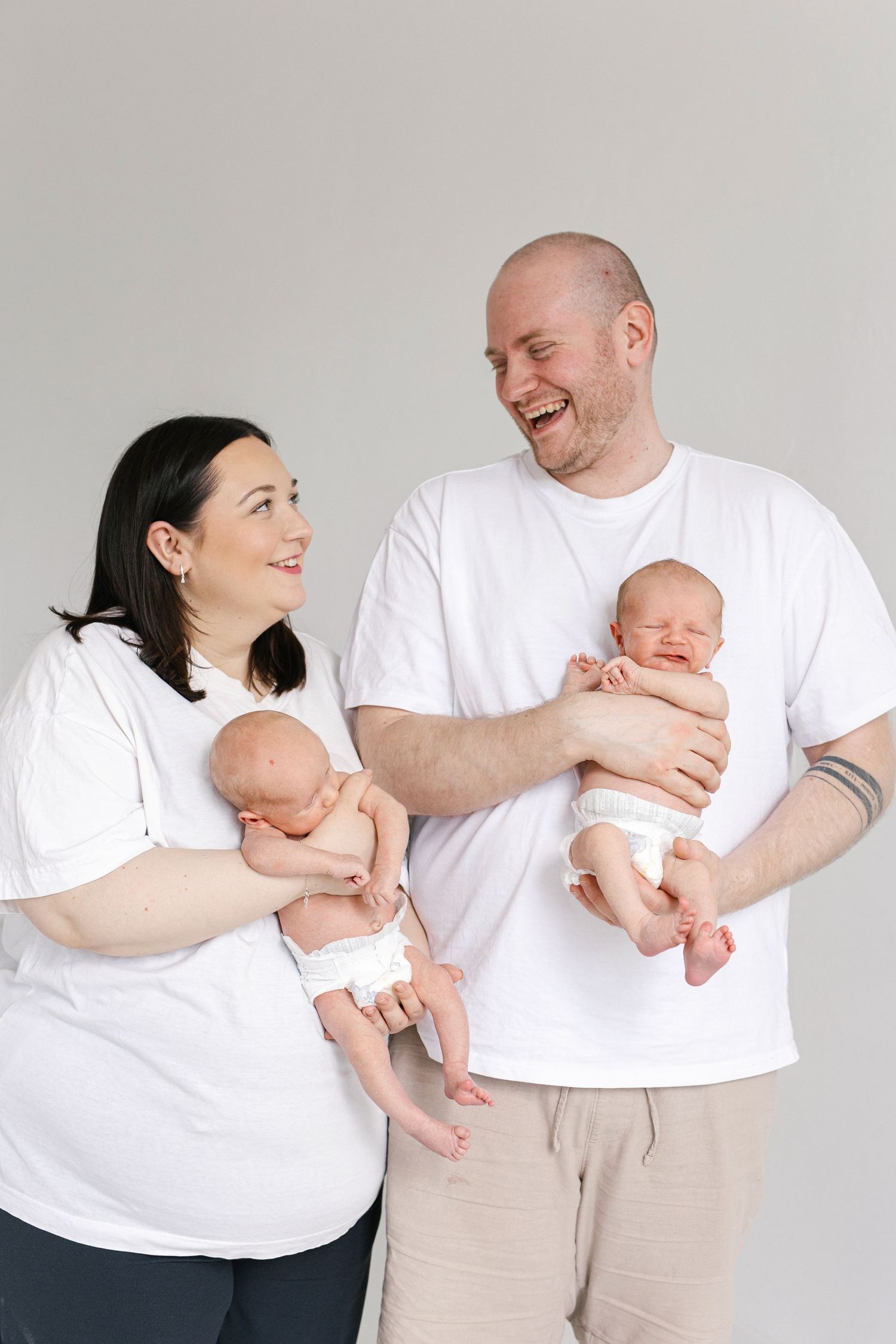 Happy parents in white t-shirts holding their newborn twins during a studio photography session.