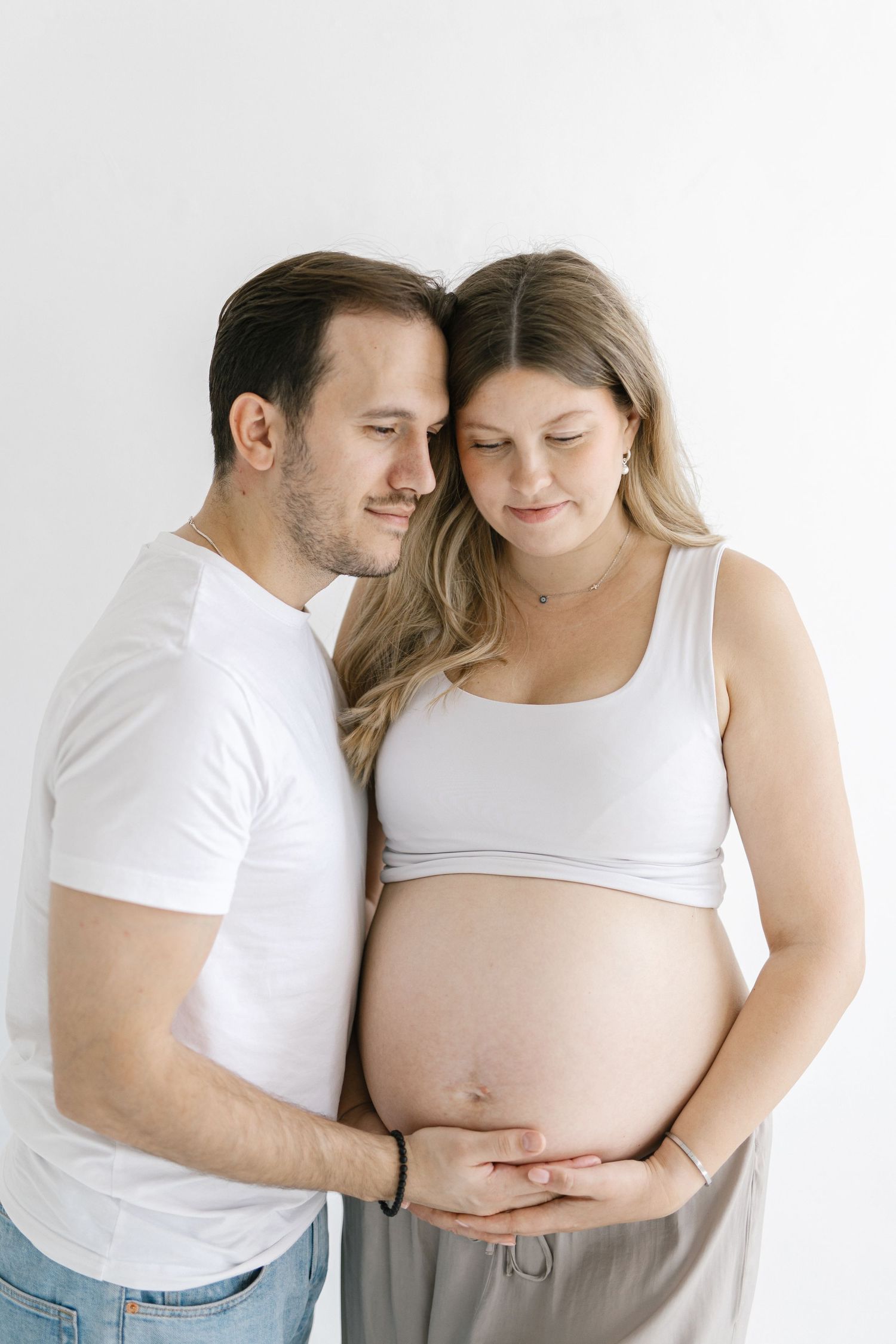 A couple tenderly holding the pregnant woman's baby bump, both looking down with gentle smiles in a bright studio.