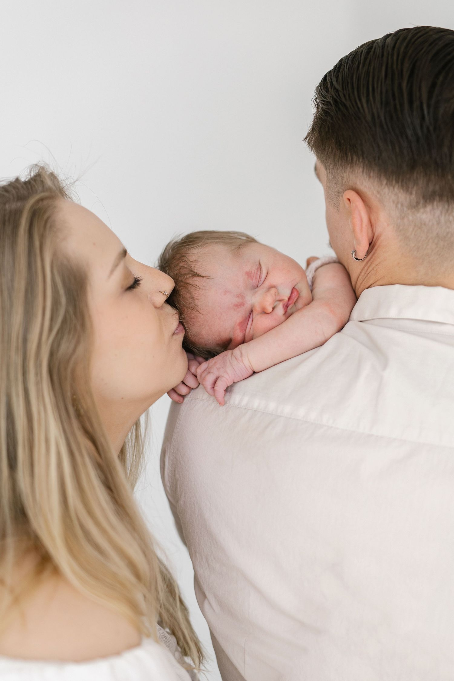 Parents kissing and holding their newborn baby against a white background in a tender family portrait.