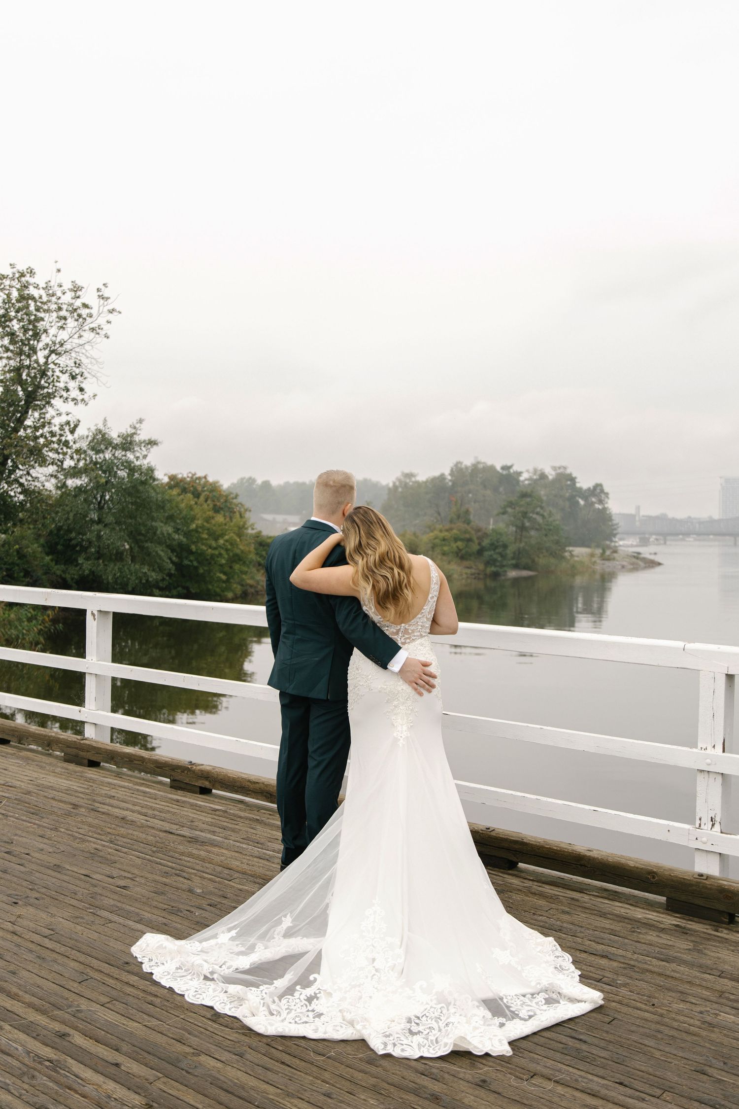 A romantic wedding couple embraces on a misty wooden dock overlooking a serene lake.