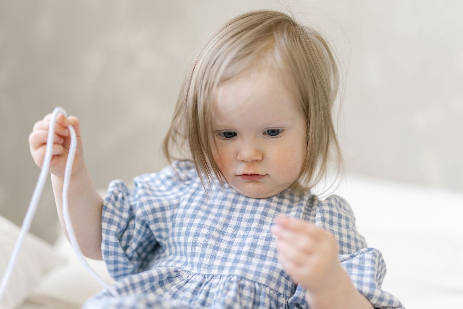 Toddler girl in blue gingham dress playing with white ribbon, looking down with concentration.