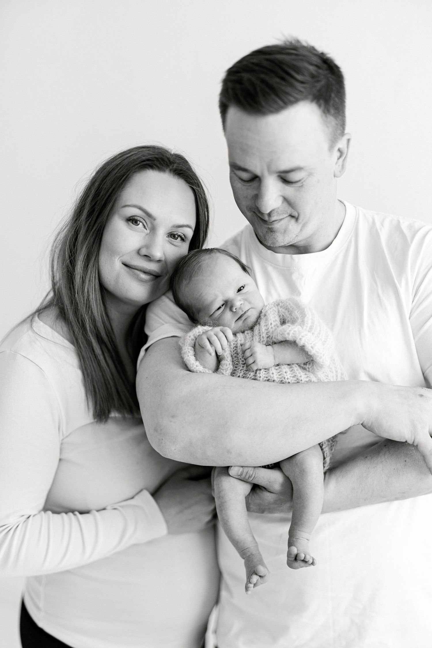 Black and white newborn family portrait of parents lovingly holding their sleeping baby in a studio setting.