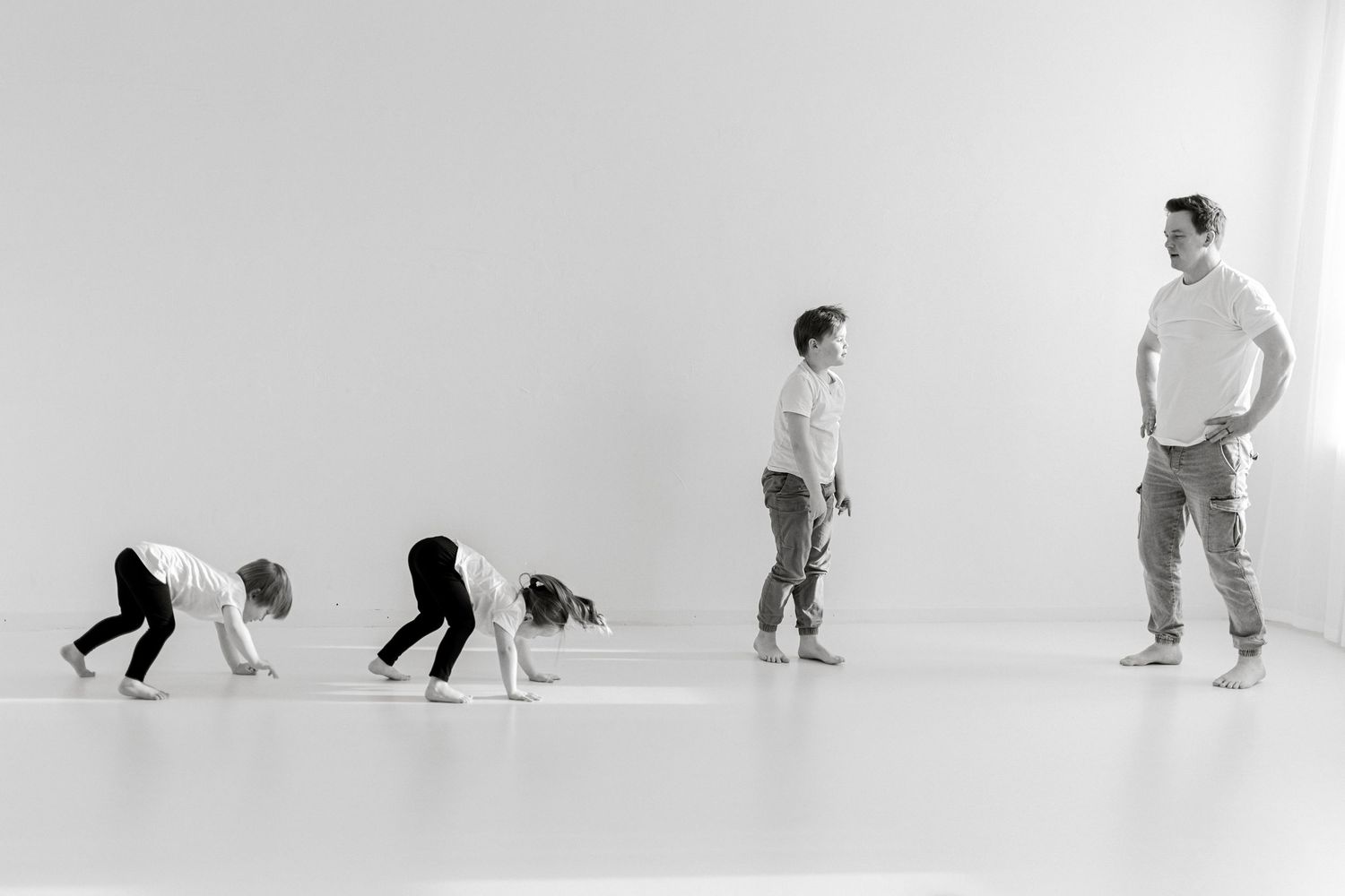 Black and white photo of father watching three children in various stages of standing up in white studio.