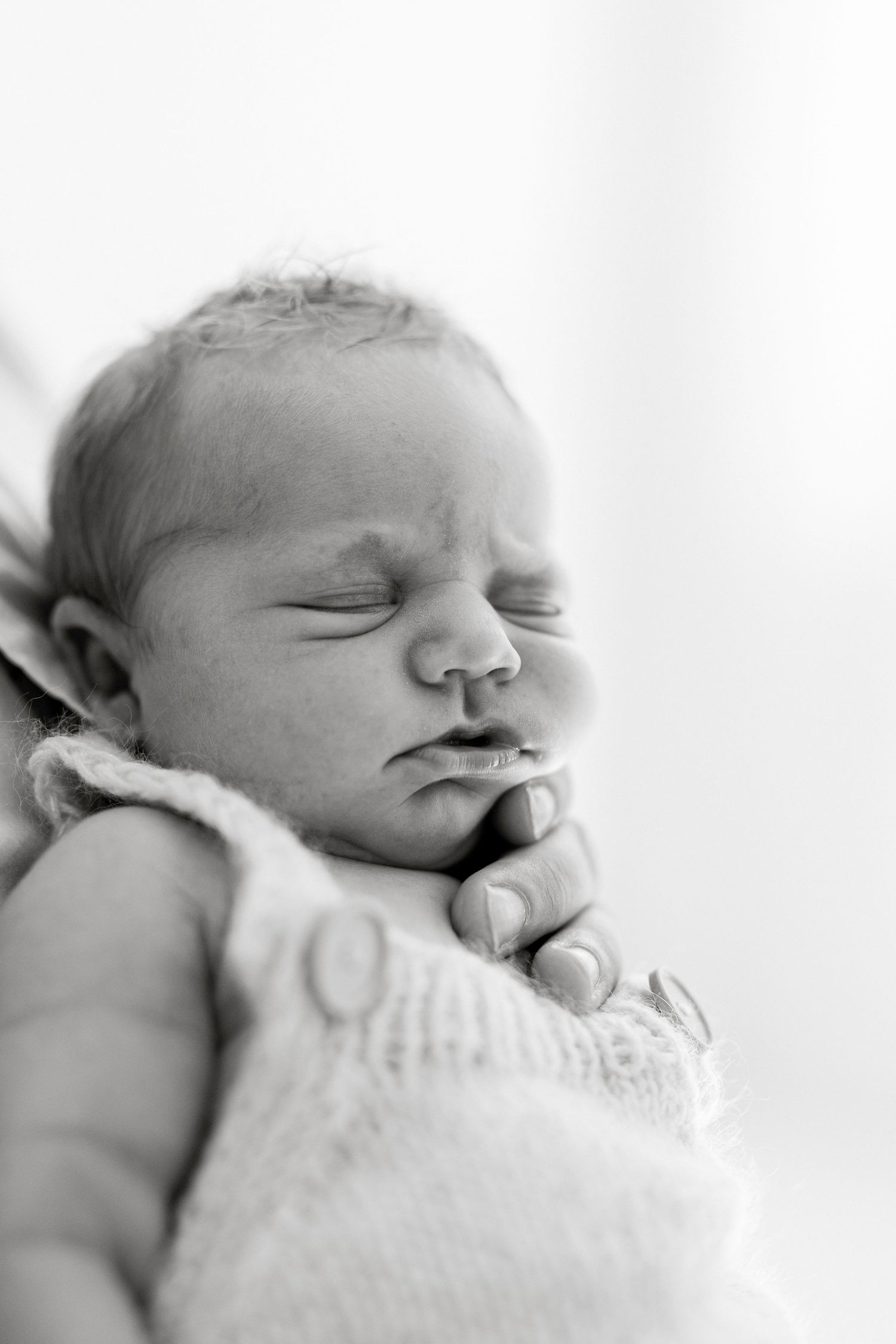 Black and white photo of a sleeping newborn baby held gently in an adult's hand, wearing a knit outfit.