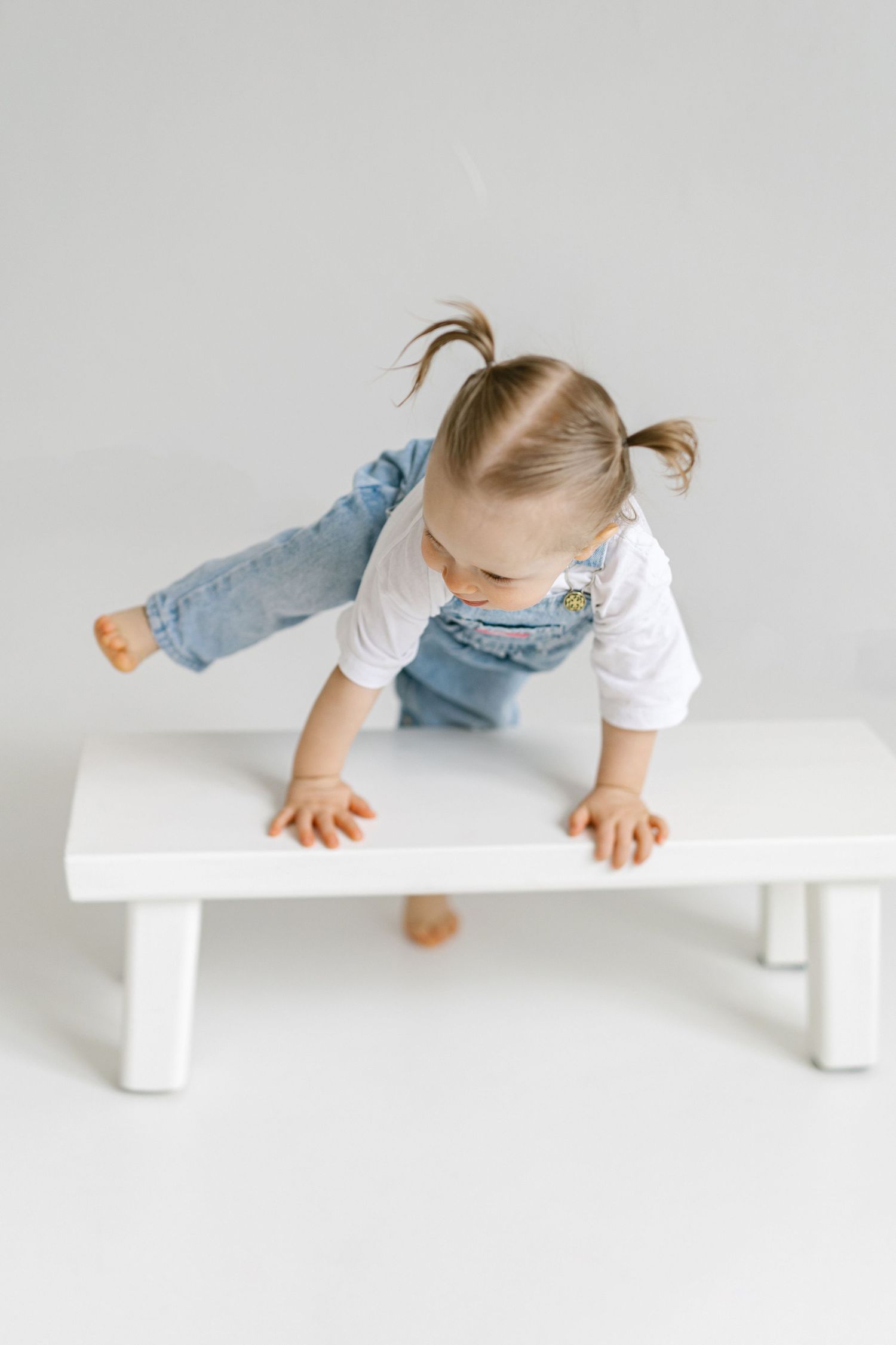 Toddler girl in denim overalls and pigtails leaning over a white wooden bench on a light gray background.