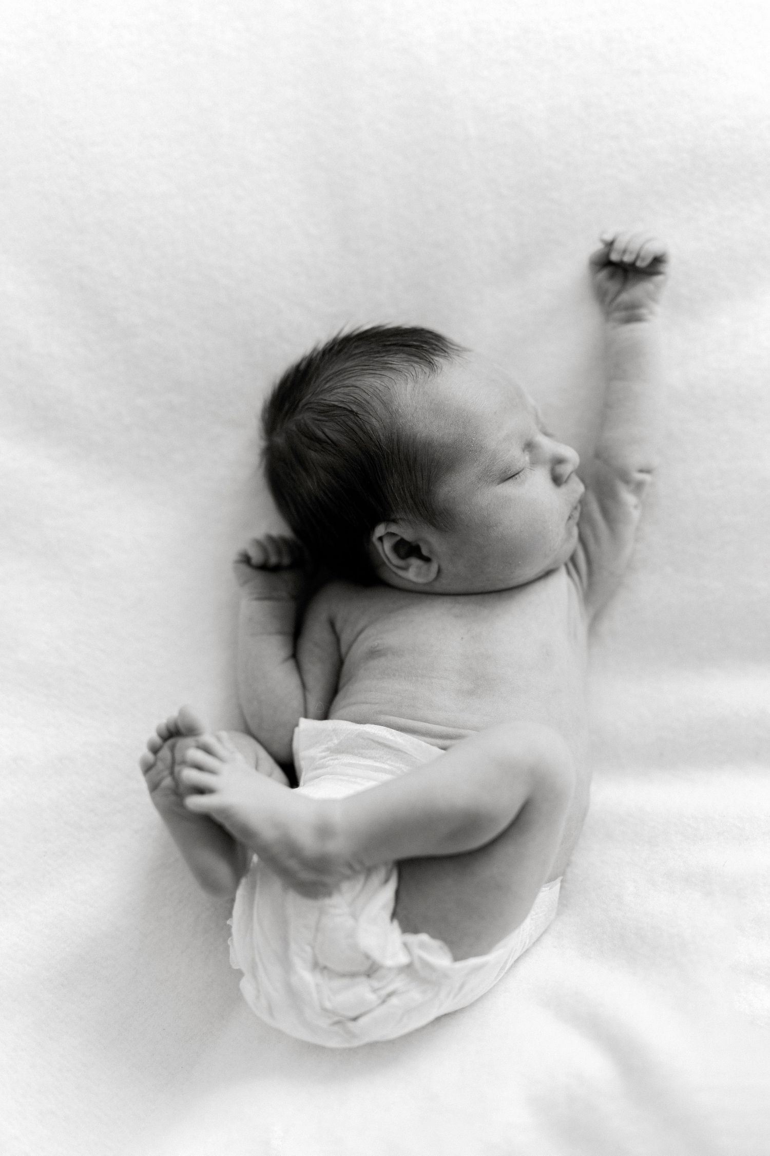 Black and white photo of a sleeping newborn baby in a diaper, stretching one arm up on a white blanket.
