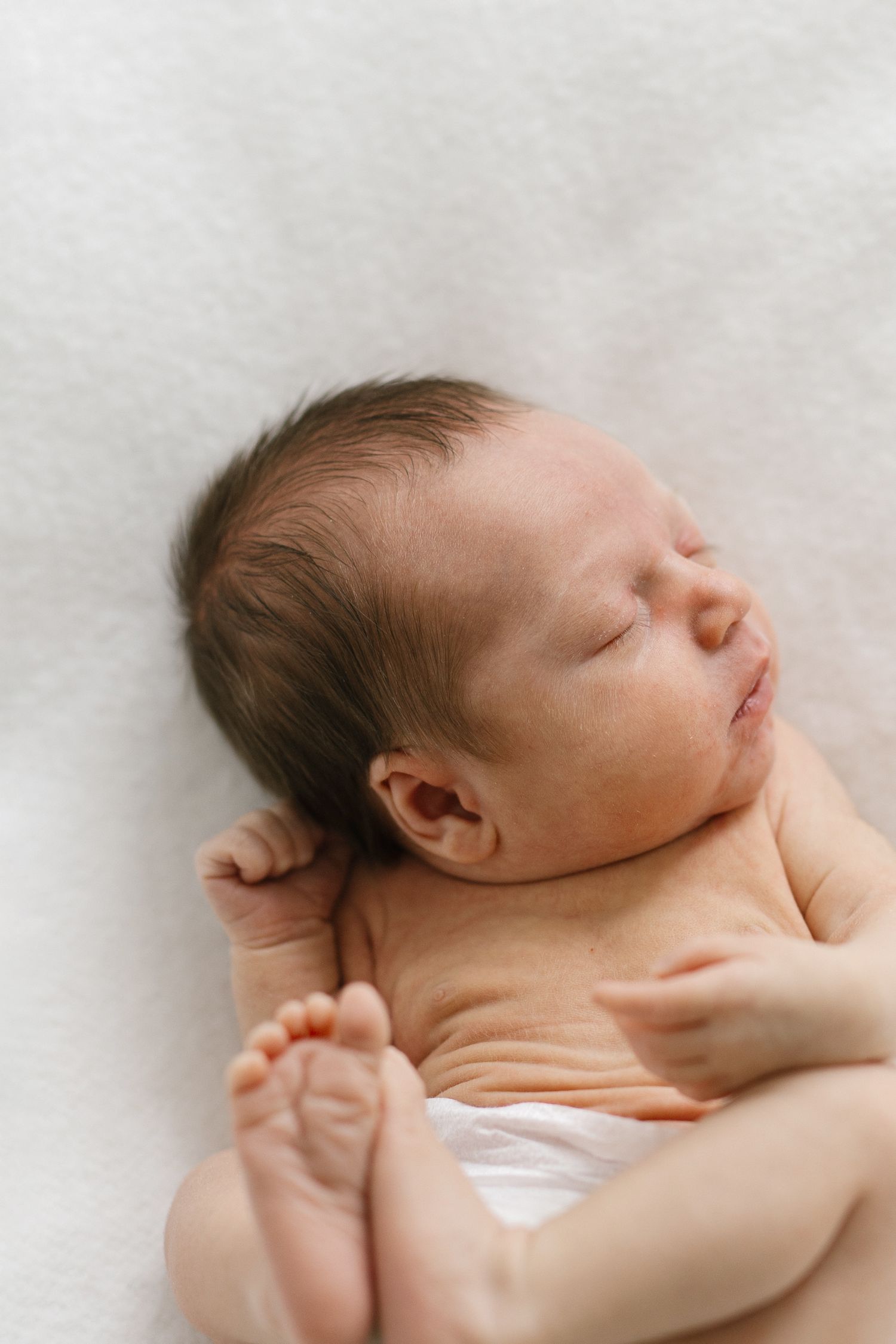 Peaceful sleeping newborn baby with dark hair lying on white blanket, tiny fists curled and feet visible.