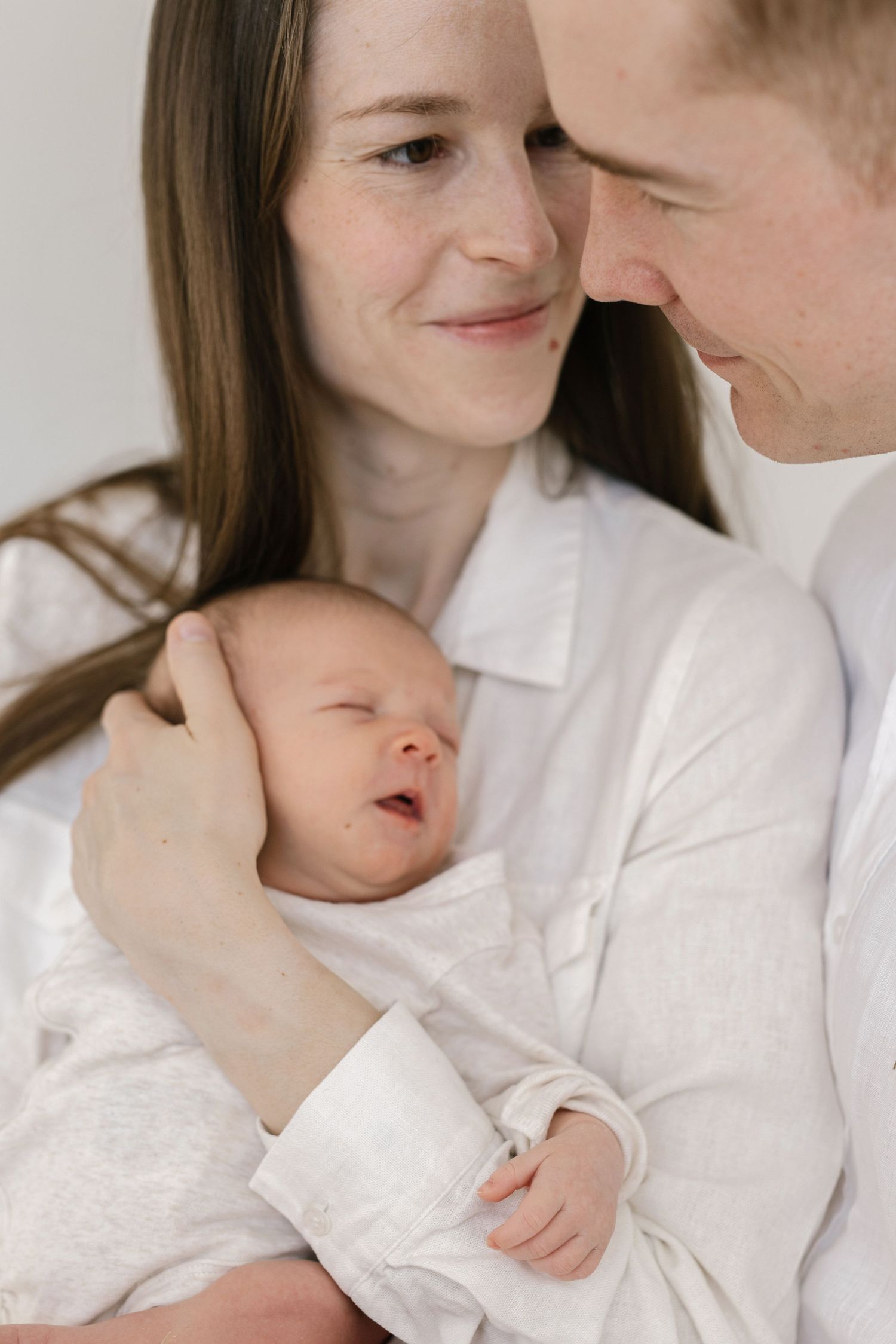 Intimate family portrait series with newborn baby in all-white setting.