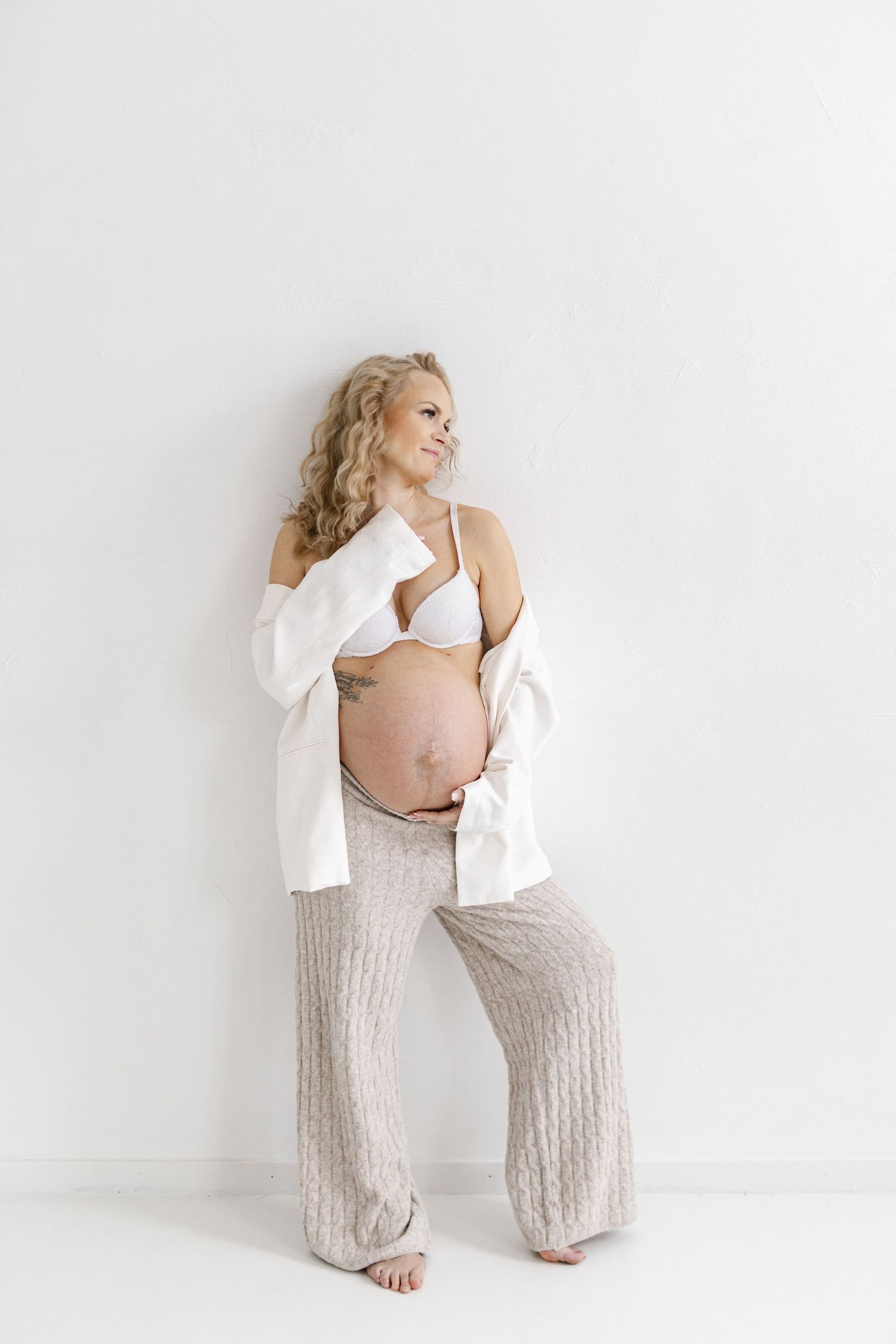 Maternity portrait with subject wearing white cardigan and beige knit pants against white background.