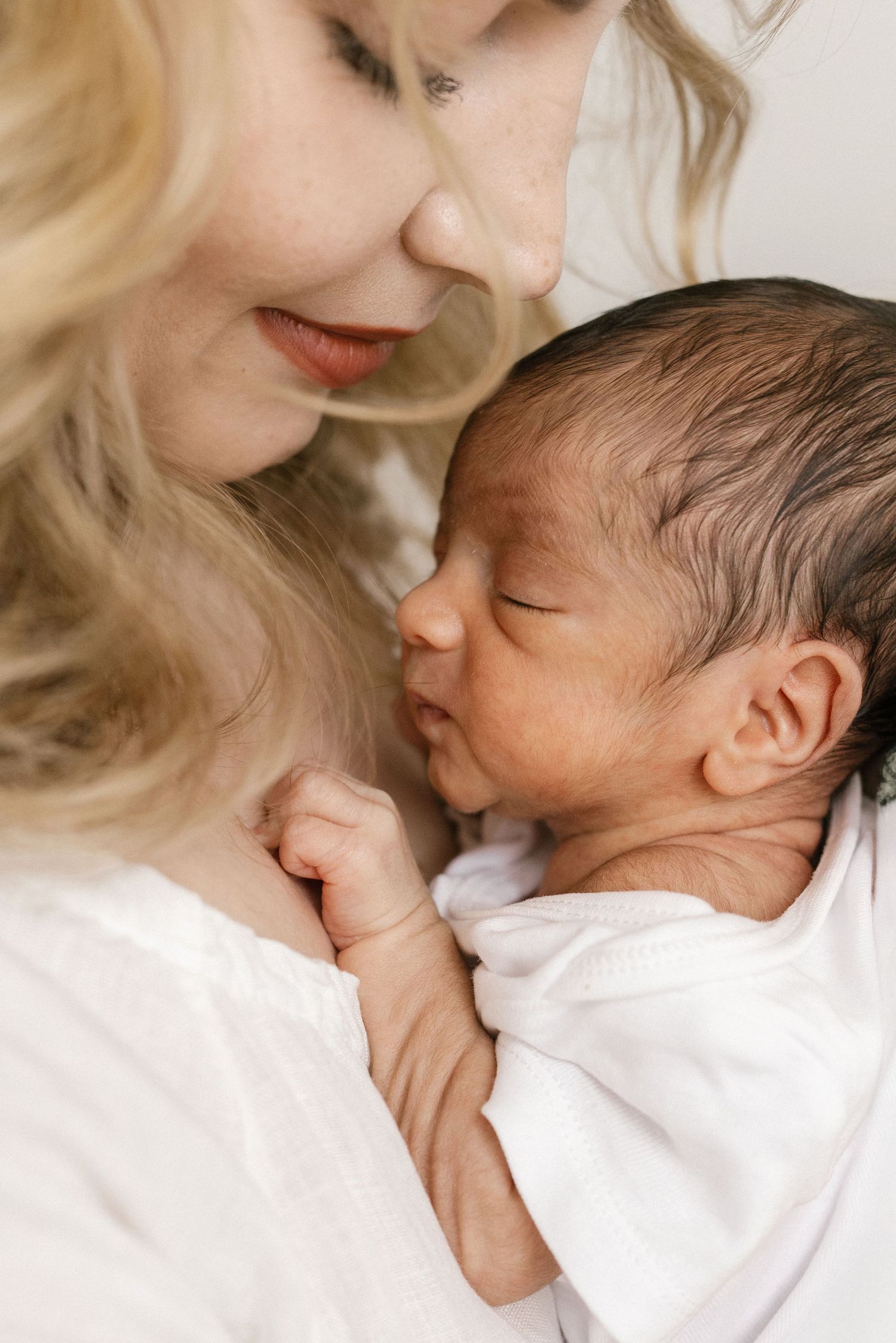 A blonde woman in white gently holds a sleeping newborn baby wrapped in a white blanket close to her chest.