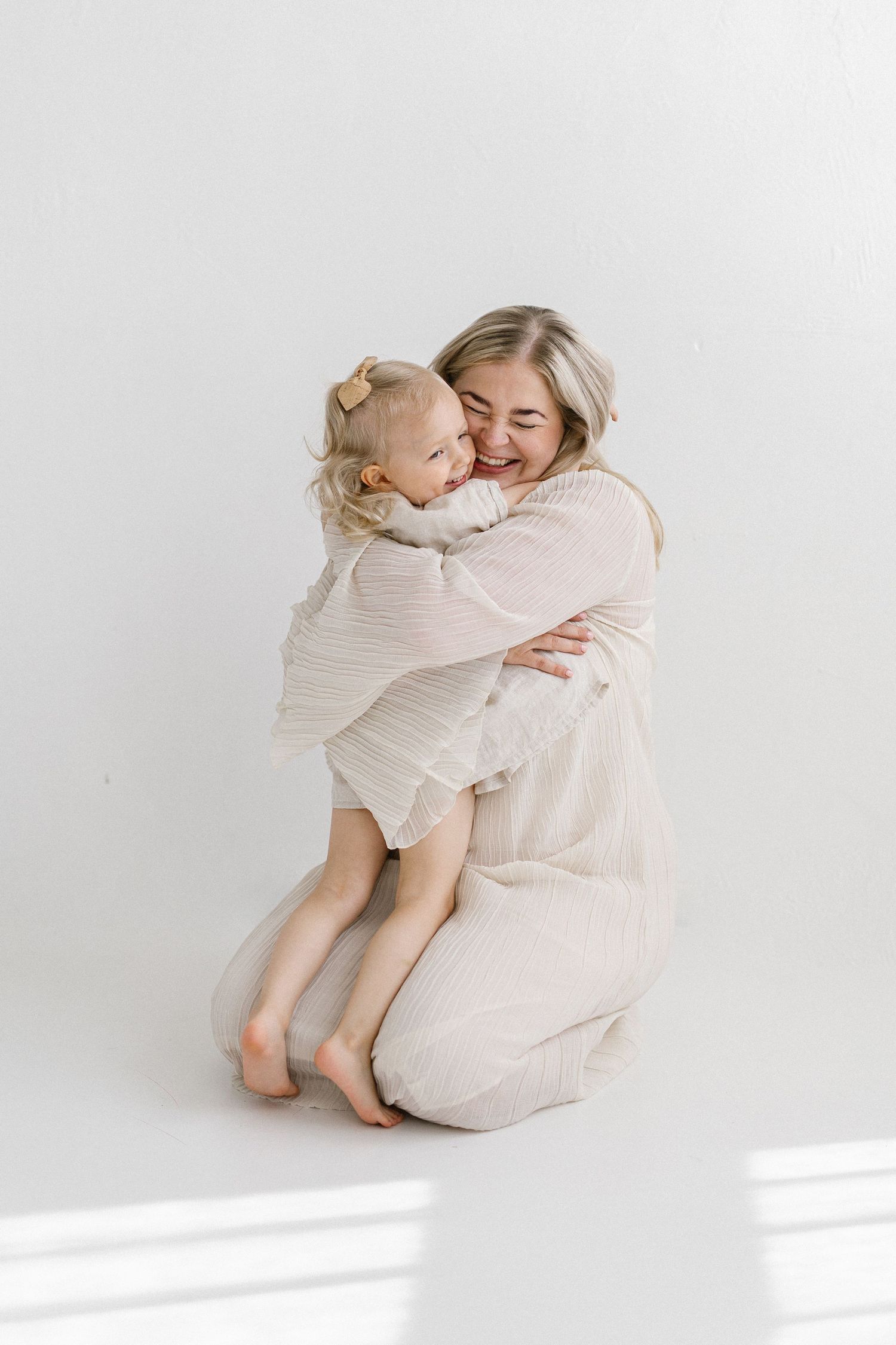 A mother in a beige dress kneels hugging her smiling toddler daughter, both wearing neutral tones against a white background.