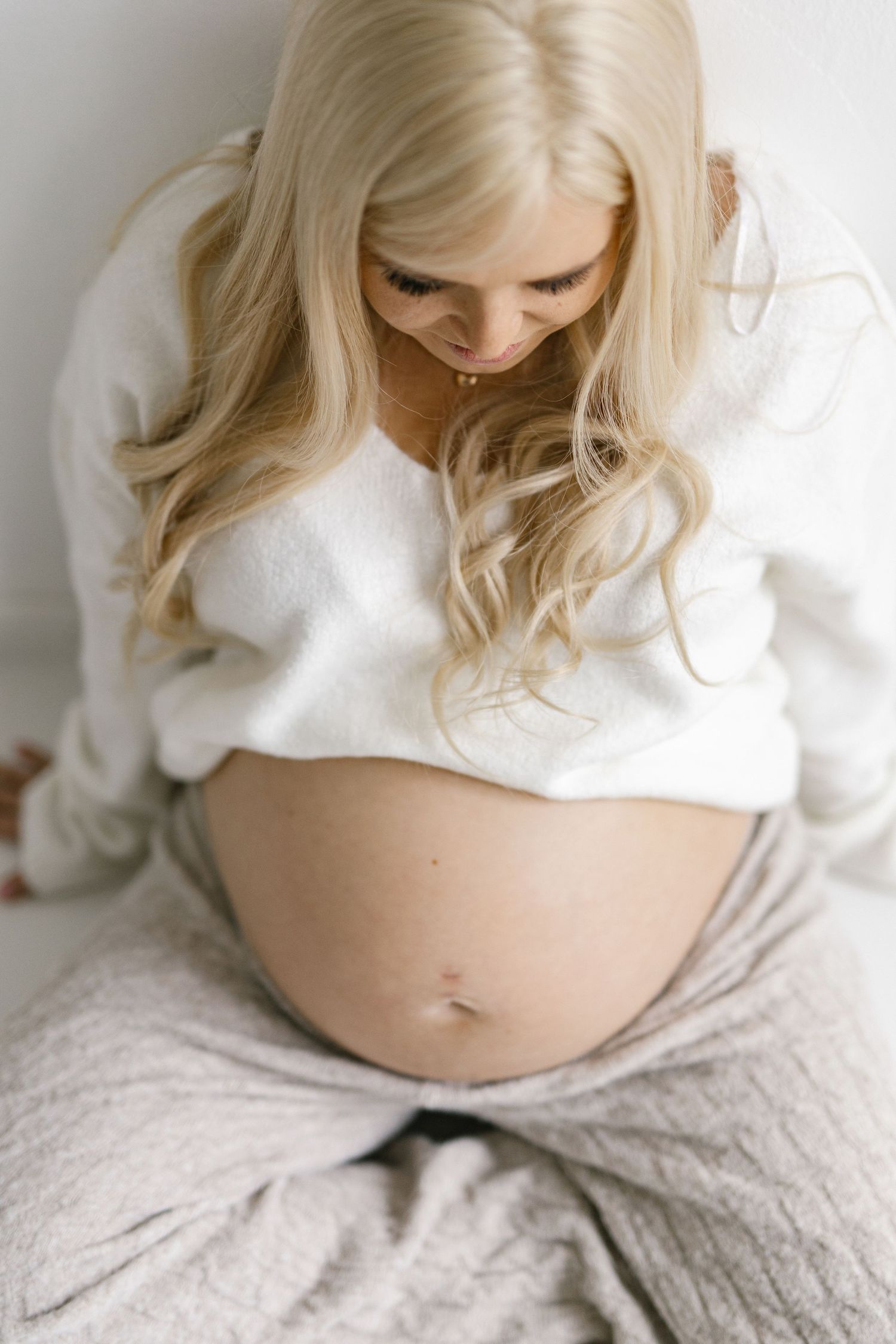 Pregnant woman with long blonde hair in white sweater looking down at her bare baby bump while sitting.