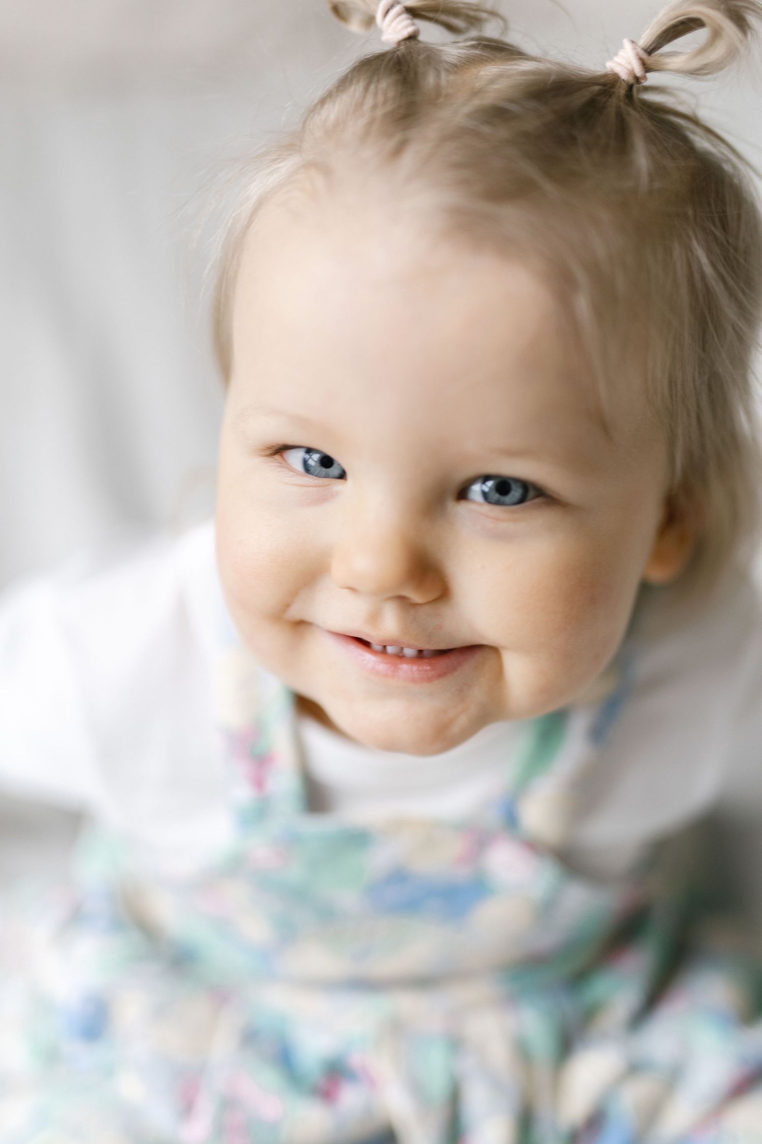 Smiling baby girl with blue eyes and pigtails wearing a floral dress, looking up with a joyful expression.