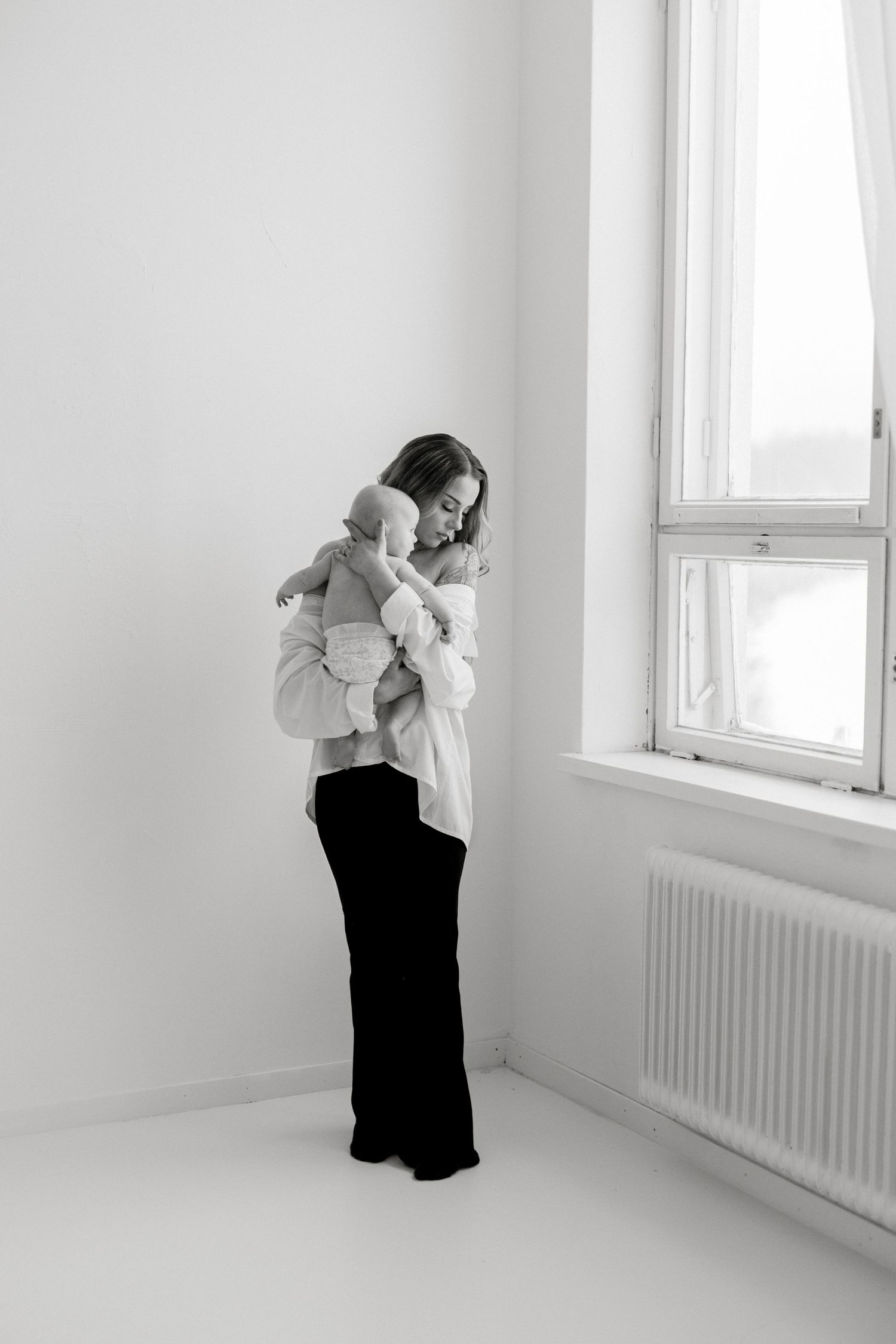 Black and white photo of a mother tenderly holding her baby near a bright window in a minimalist white room.