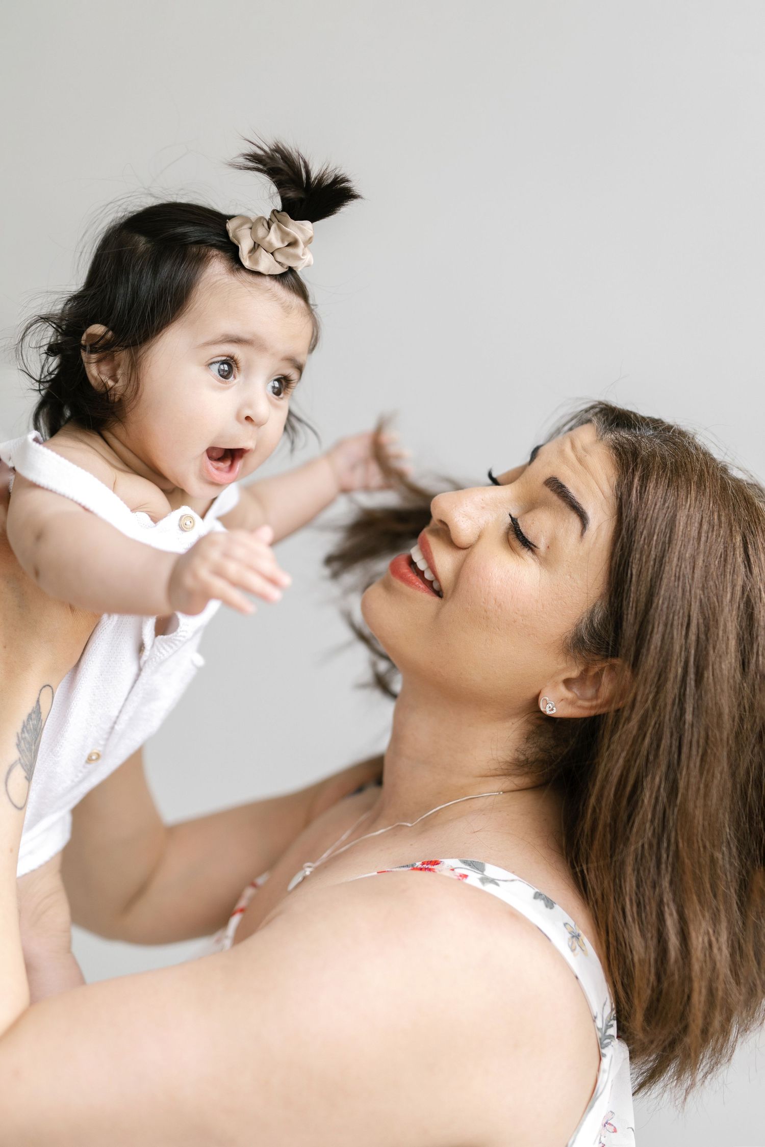 Joyful mother lifting her surprised baby girl with dark hair and beige scrunchie against gray background.