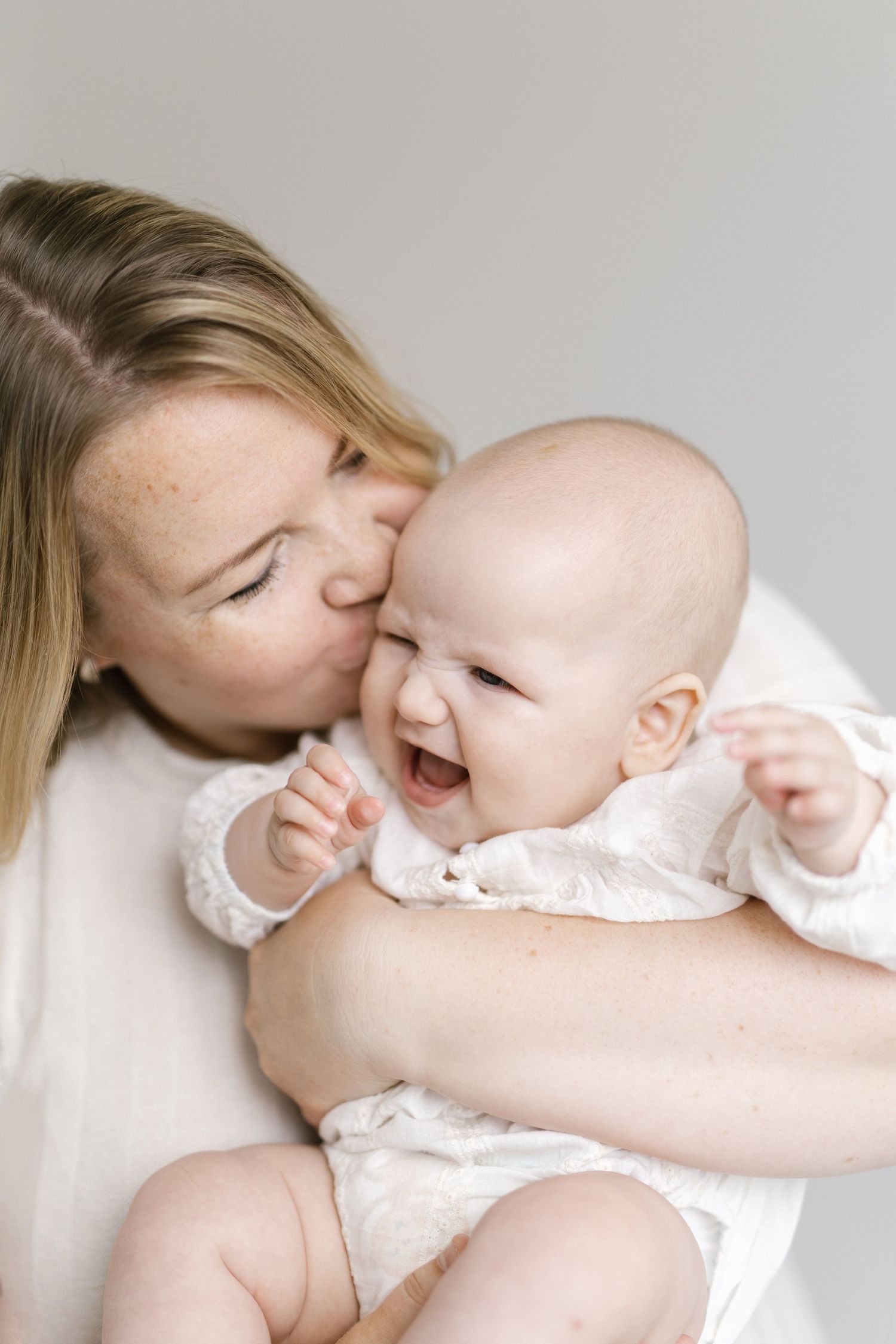 A mother with freckles kisses her laughing baby dressed in white, sharing a joyful moment together.