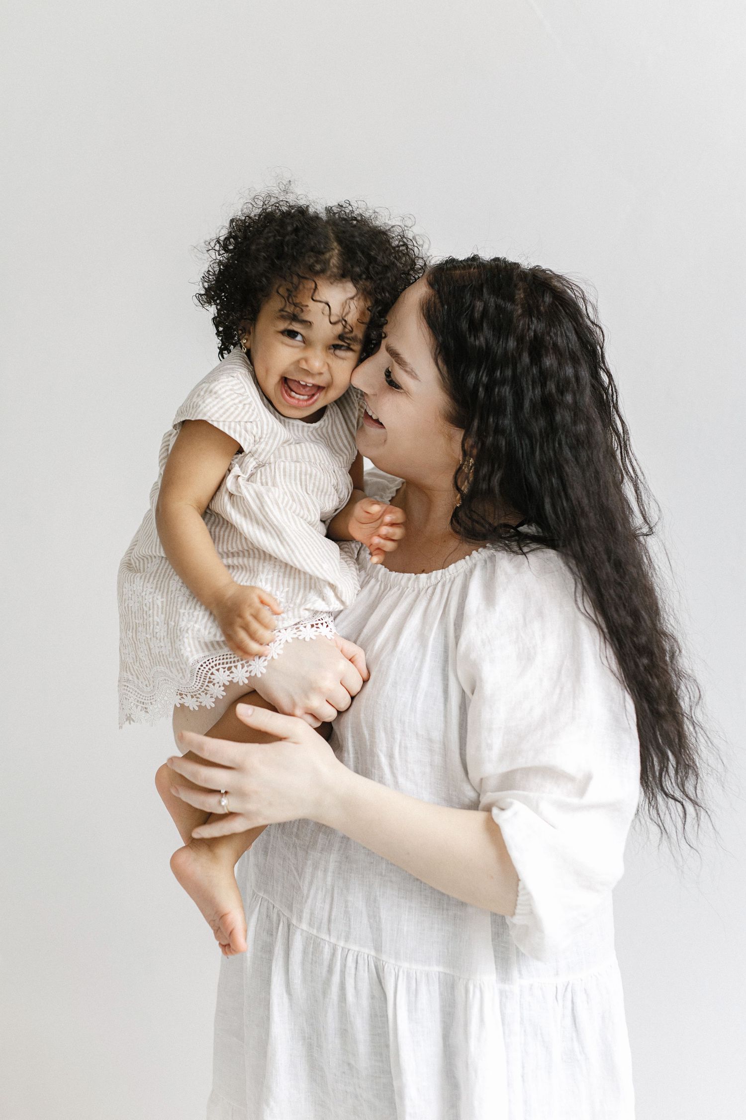 A young woman in a white dress lovingly holds a laughing toddler girl with curly hair against a white background.