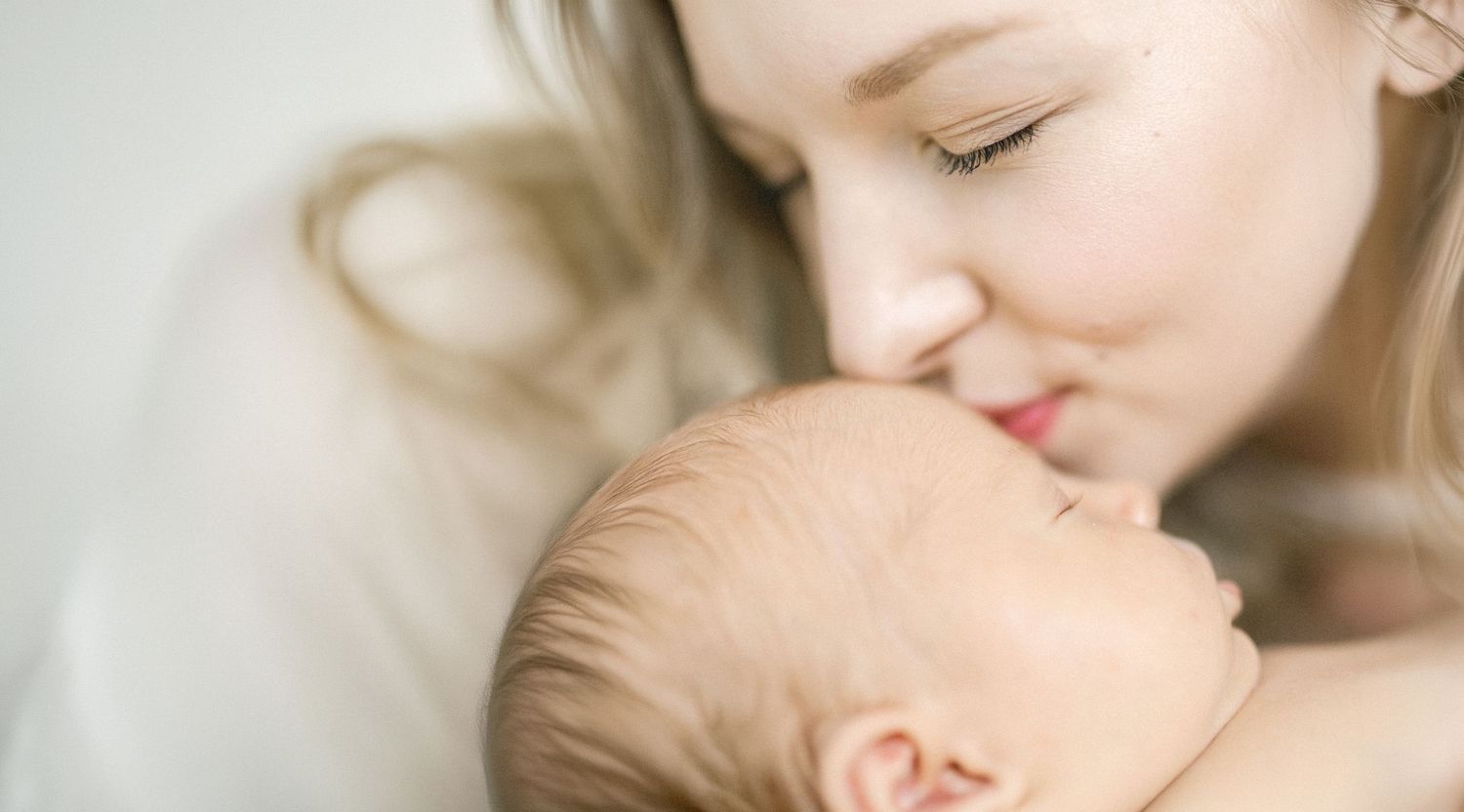 A tender moment as a young mother gently nuzzles her sleeping newborn baby, captured in soft natural light.