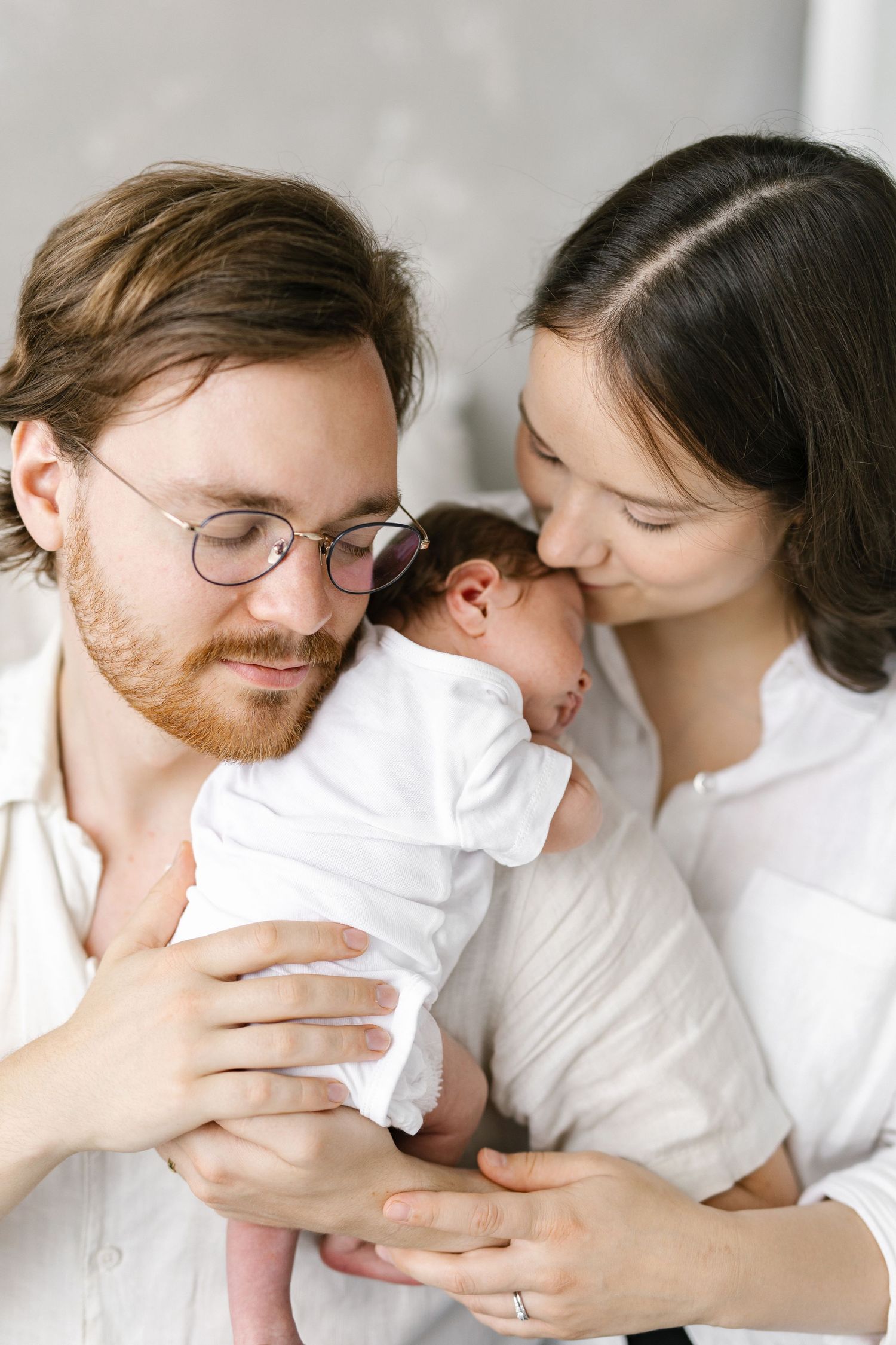 Young parents with red-bearded father in glasses and dark-haired mother tenderly holding their sleeping newborn baby.