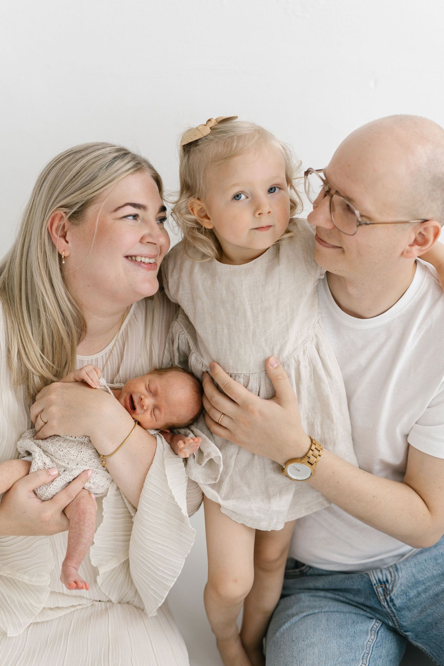 A happy family of four with a newborn baby, toddler girl, mother, and bald father posing together in soft neutral tones.