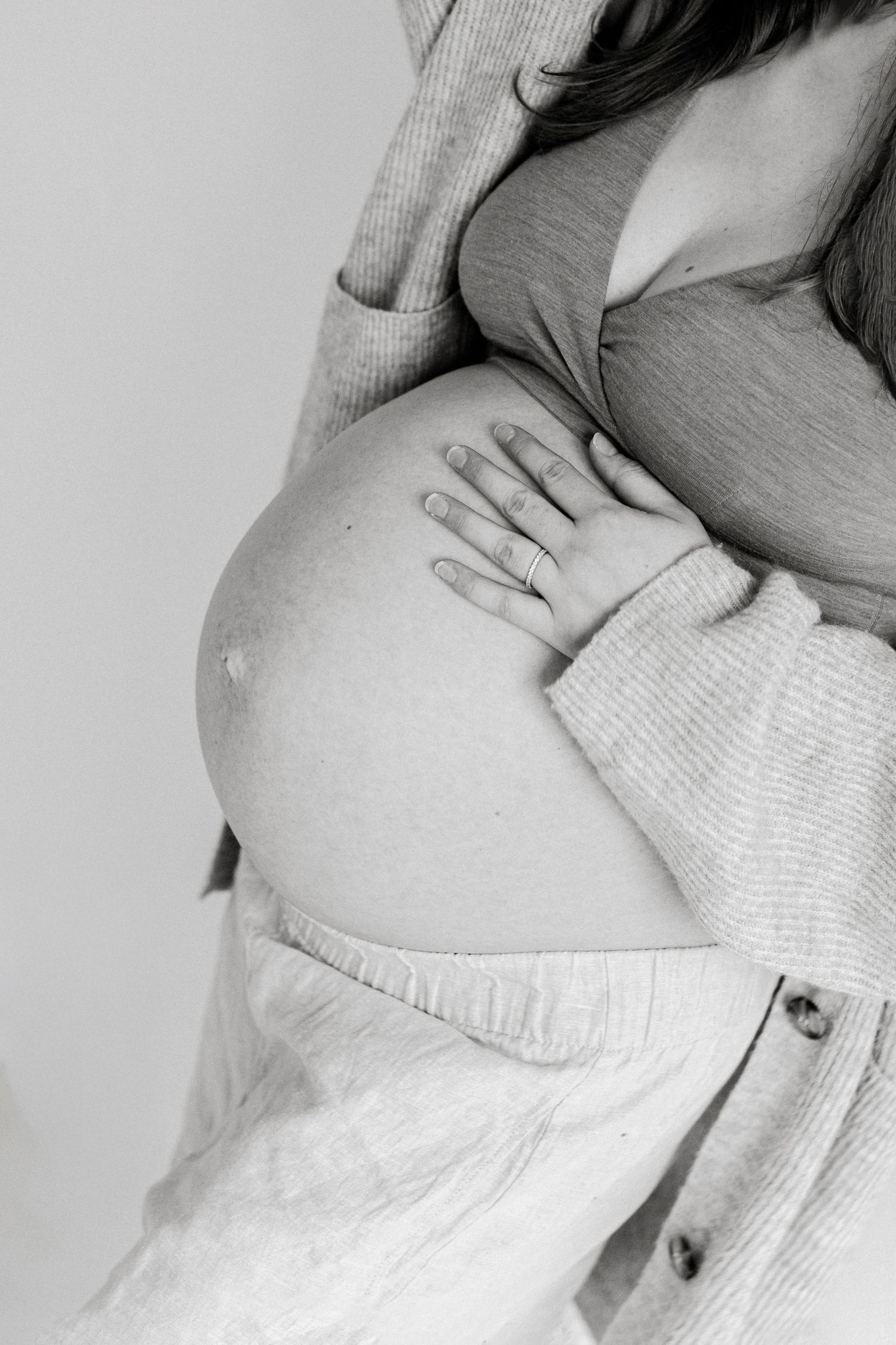 Black and white maternity photo of a pregnant woman in a cardigan and linen pants, her hand with a ring resting on her bare belly.
